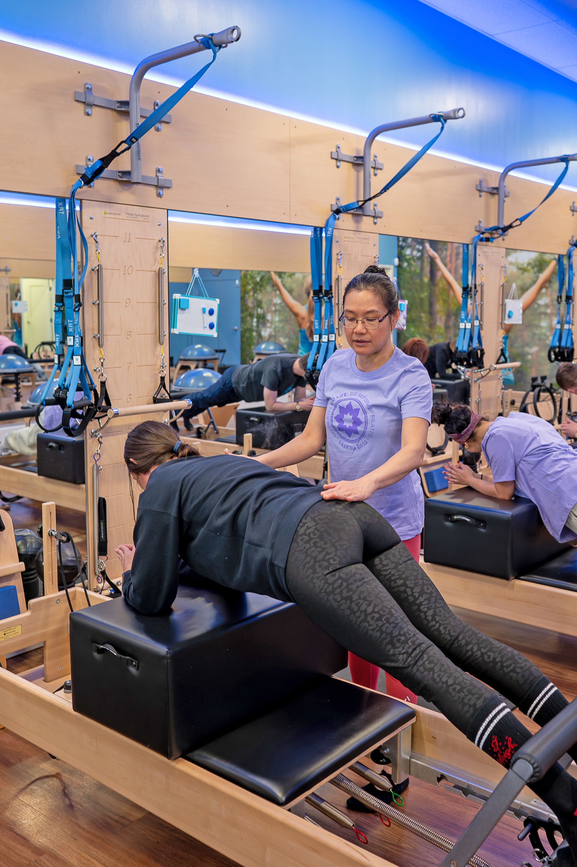 An instructor observes a client performing a plank exercise on a Pilates reformer in a studio.