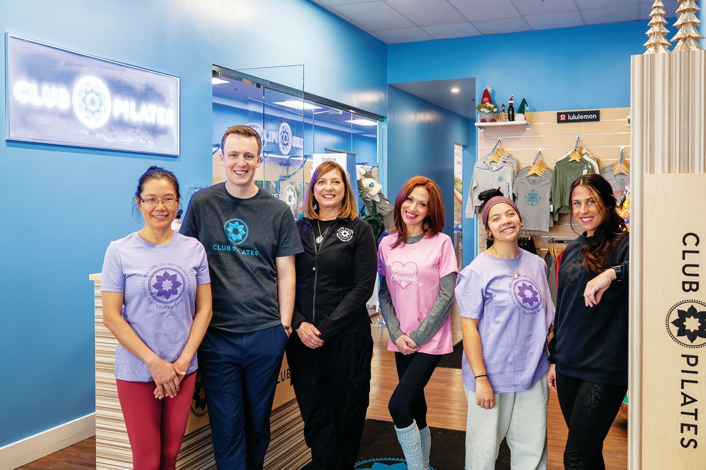 Six smiling people stand in a brightly lit Club Pilates studio, posing together in casual athletic apparel.