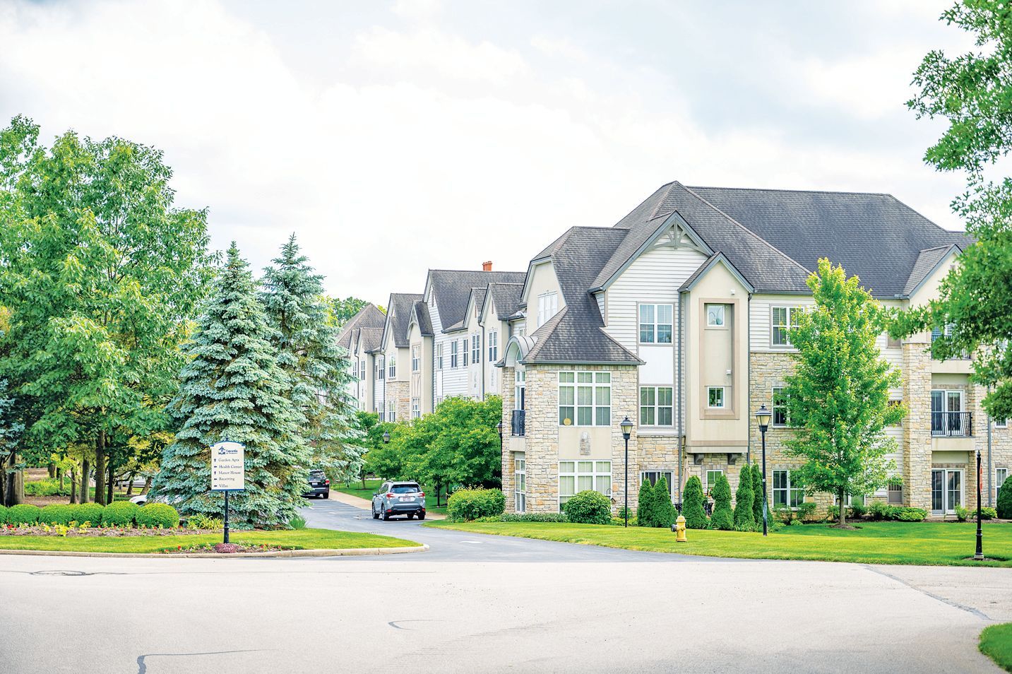 A three-story apartment complex with stone and siding exterior, surrounded by lush green trees and a paved driveway.