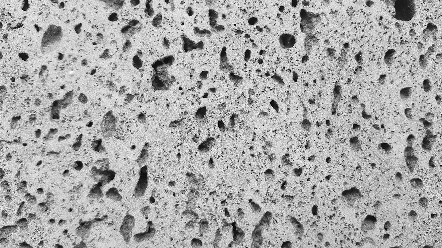 A close-up, black-and-white texture of porous, grey volcanic rock or concrete with small, irregular holes throughout.