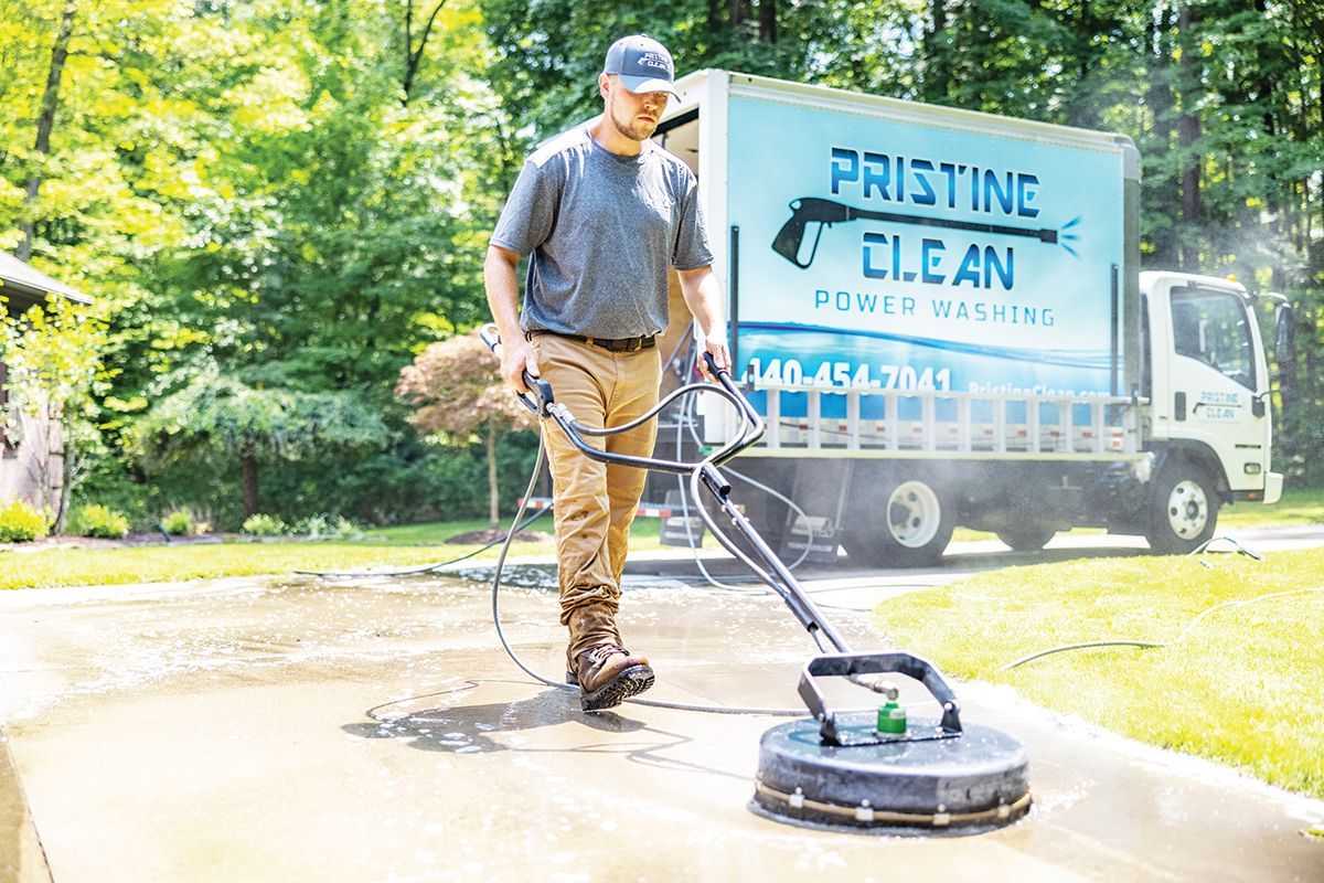A person in a gray t-shirt and tan pants pressure washing a concrete driveway near a Pristine Clean company truck.