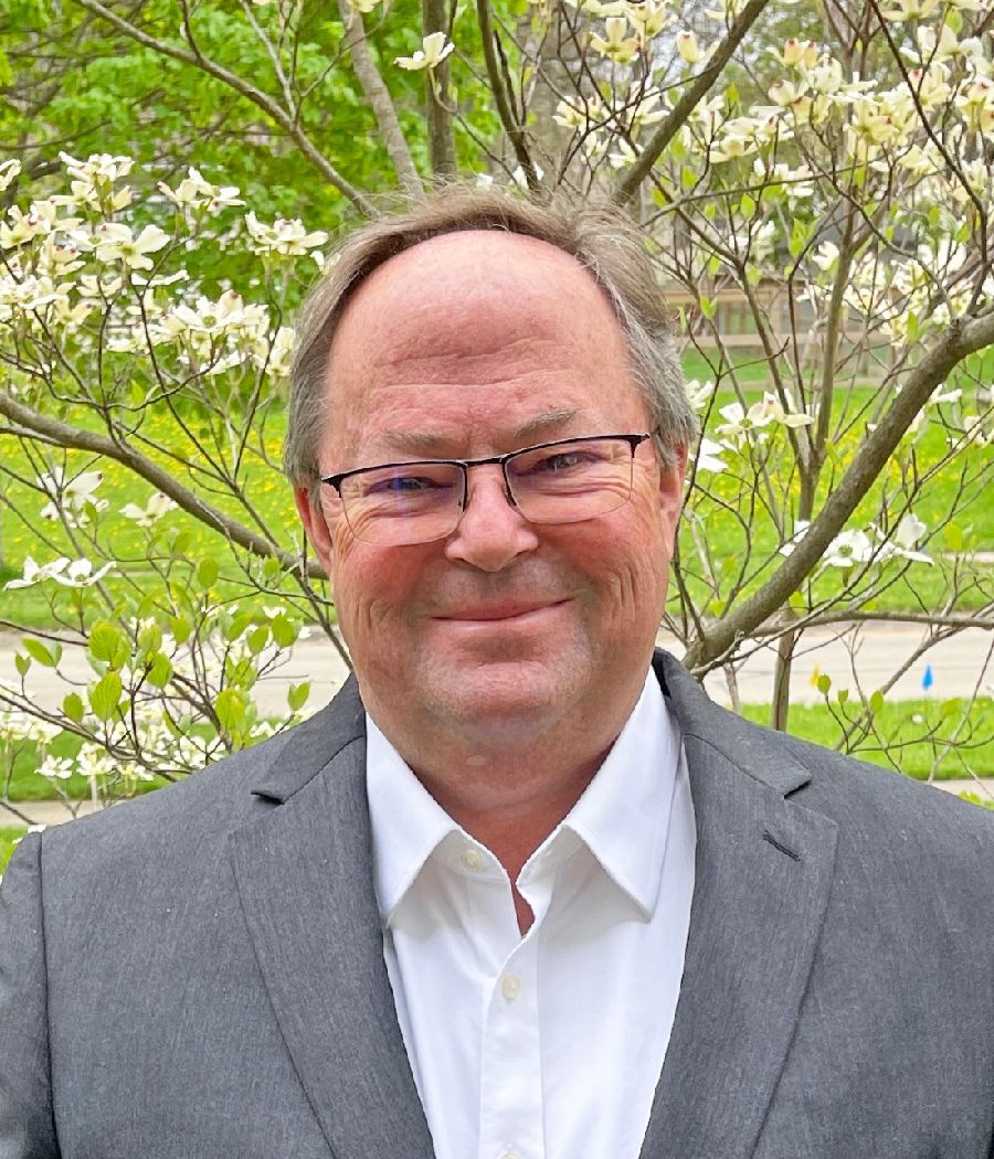 A person wearing a grey blazer and white collared shirt stands outdoors in front of a blooming dogwood tree.
