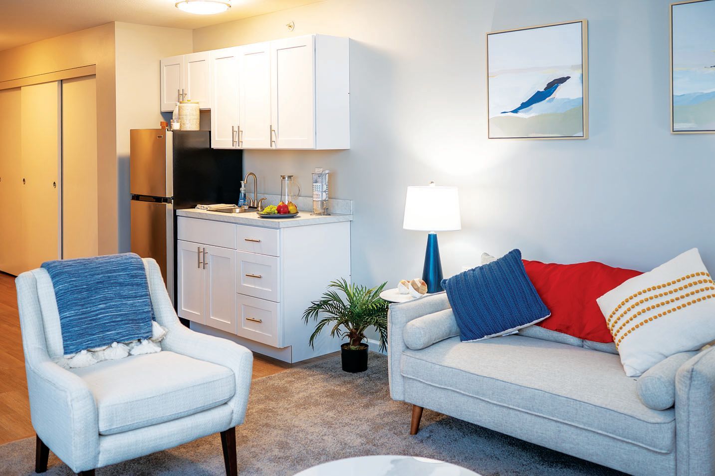 A small apartment living room featuring a light gray couch with decorative pillows, an armchair, and a kitchen area.