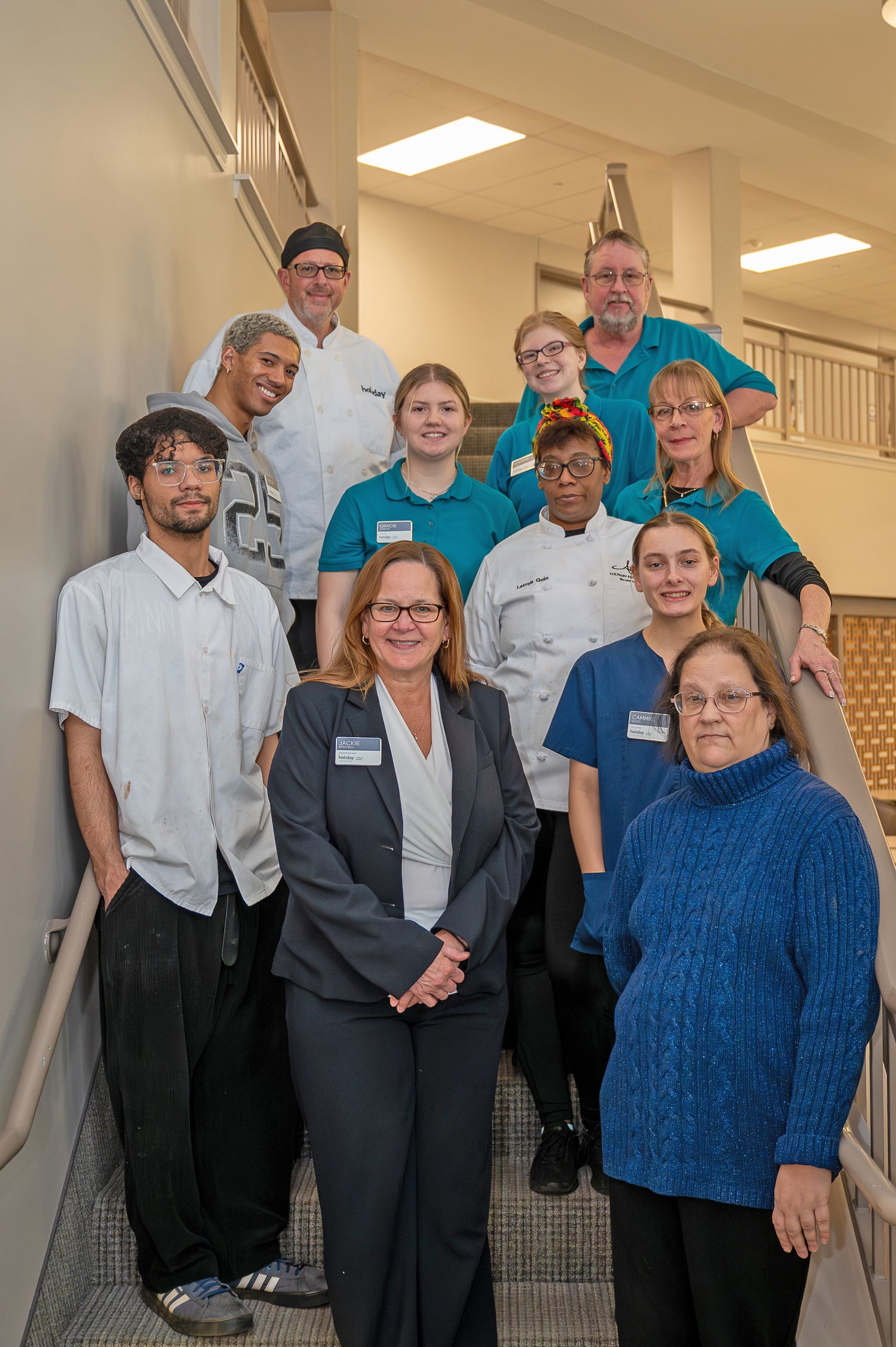 A group of people wearing varied work attire, including chef coats and blue shirts, stand together on a stairwell.