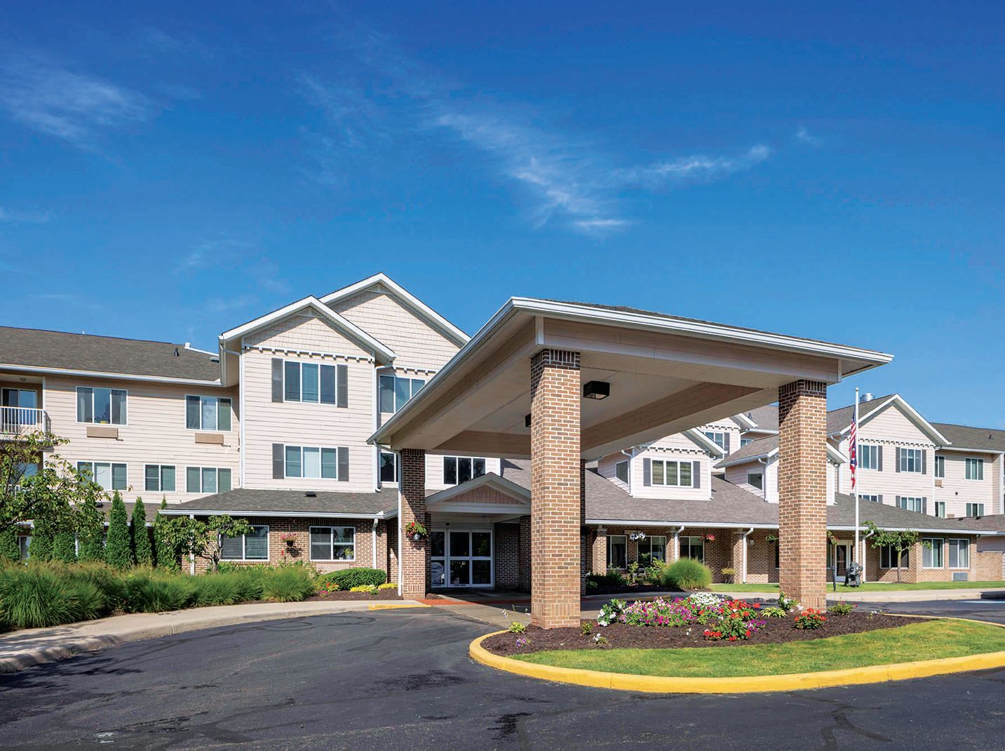 A multi-story assisted living facility with a beige siding exterior, a large entrance portico, and a paved circular drive.