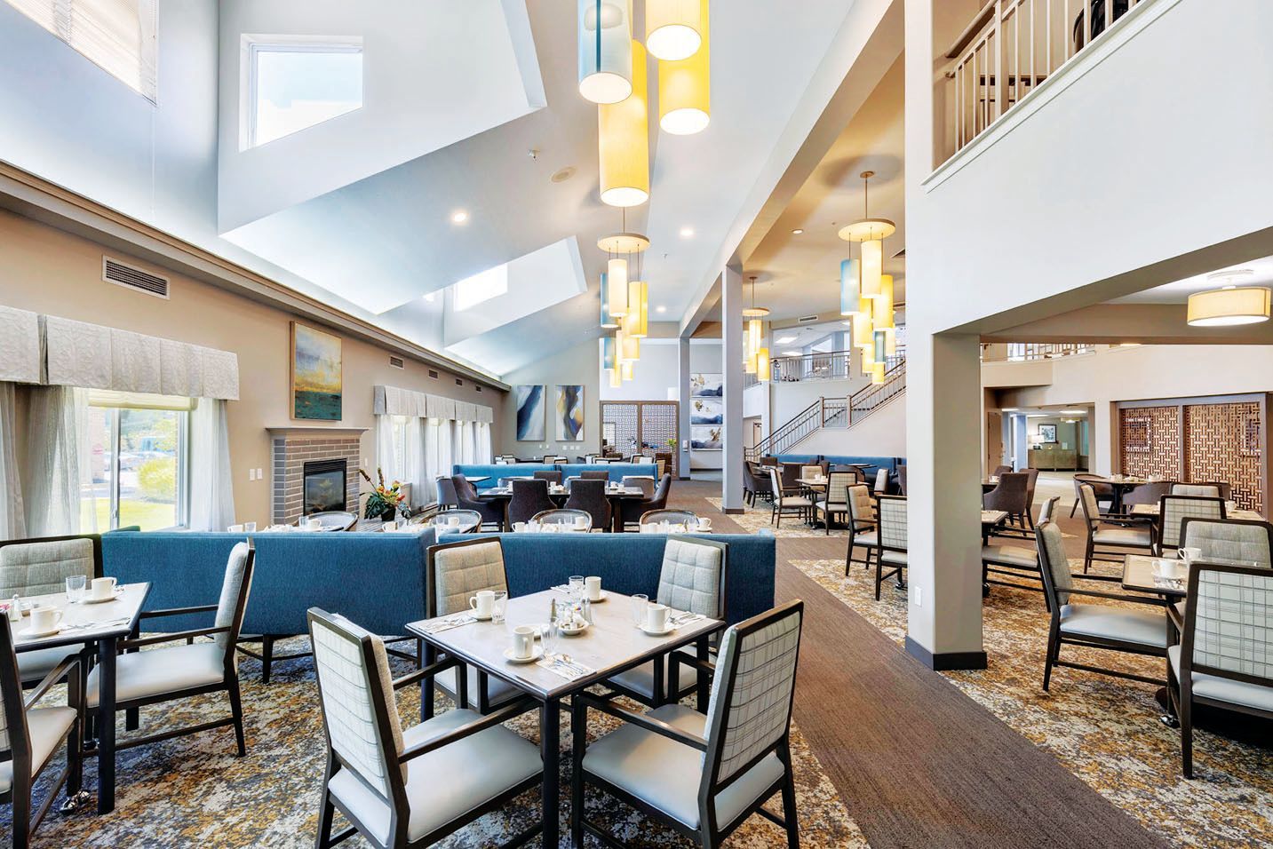 A dining room featuring square tables with chairs, blue cushioned booth seating, high vaulted ceilings, and hanging lights.
