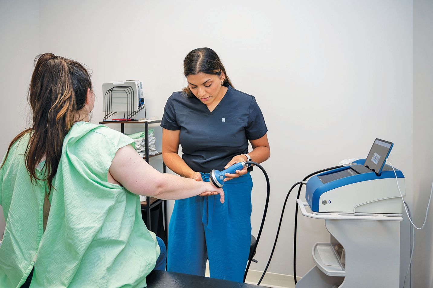 A clinician uses a handheld medical device to treat a patient's wrist in a clinic room with medical equipment nearby.