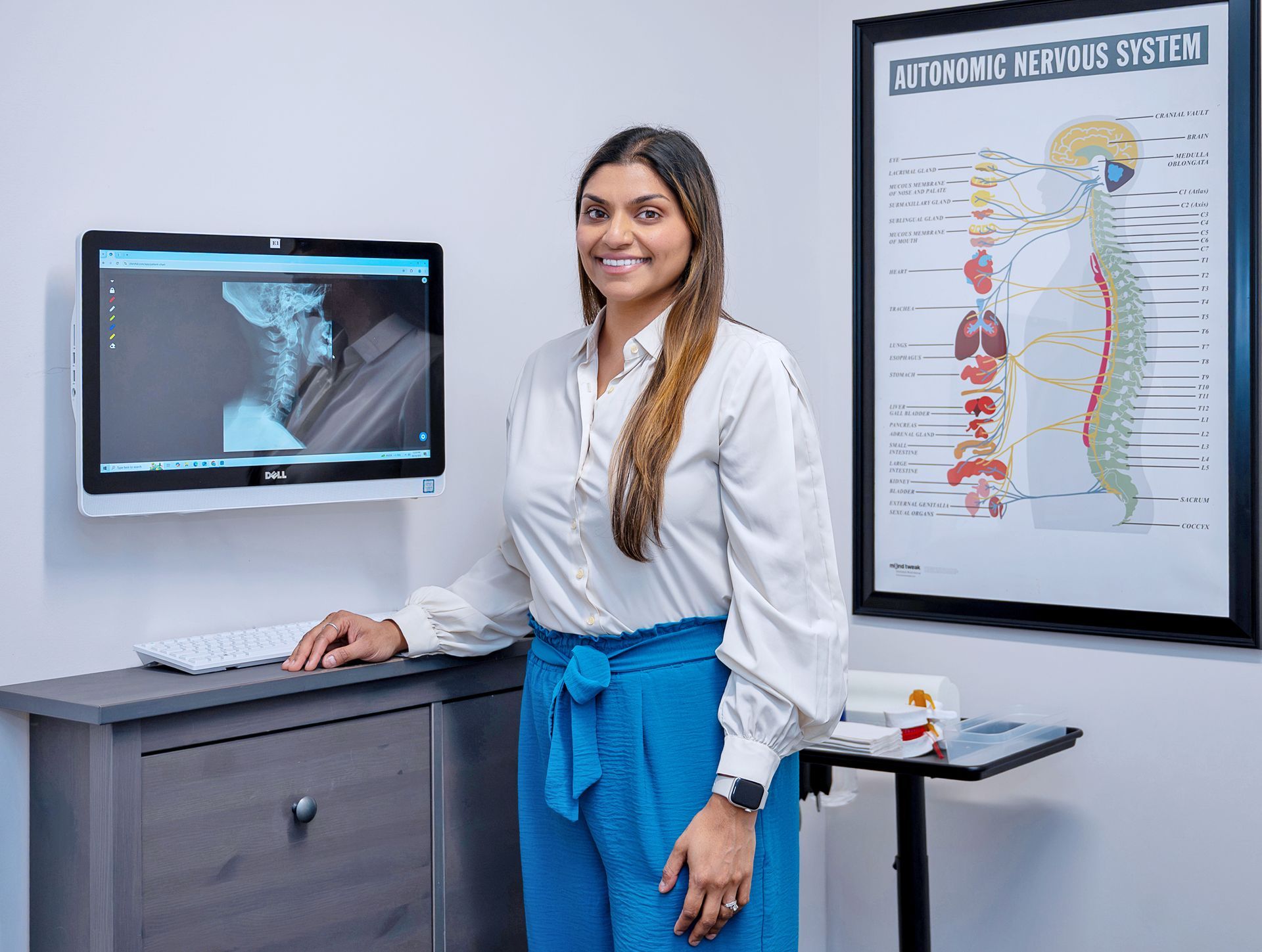 A professional smiling in a clinical office, standing next to a medical monitor and an autonomic nervous system chart.