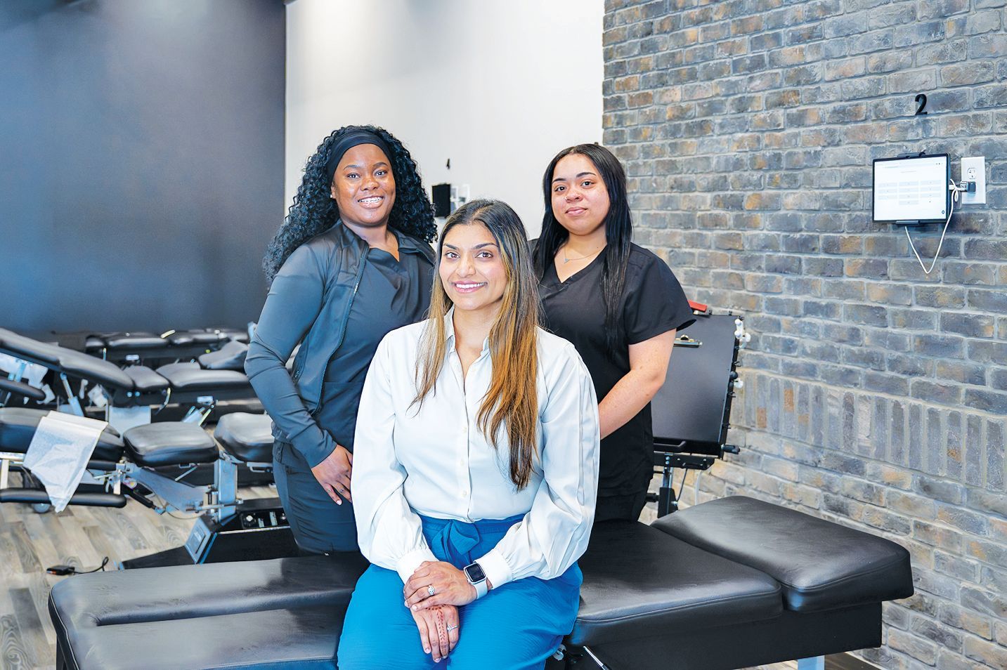 Three professionals stand in a modern clinic with treatment tables and brick walls, smiling at the camera.