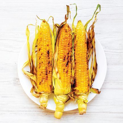 Three grilled corn on the cobs with charred kernels and husks arranged on a white plate against a light wooden background.