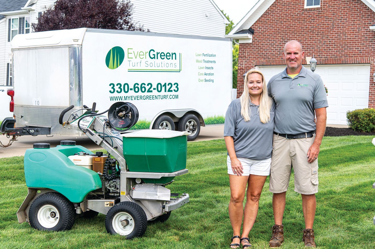 A smiling couple stands on a mowed lawn next to a green lawn care machine and a white trailer labeled EverGreen.