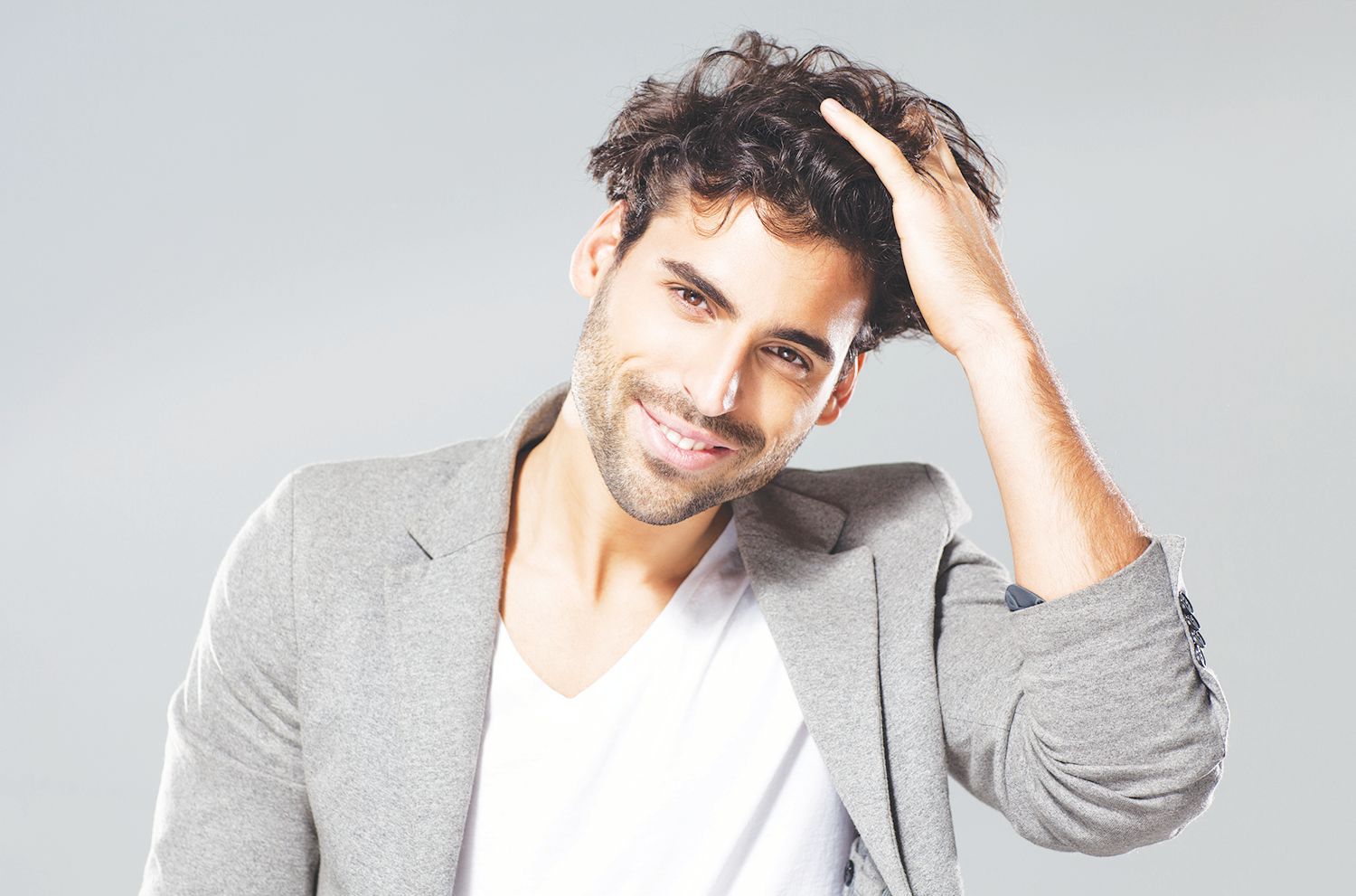 Smiling person in a grey blazer and white shirt, touching their dark, curly hair against a light grey background.