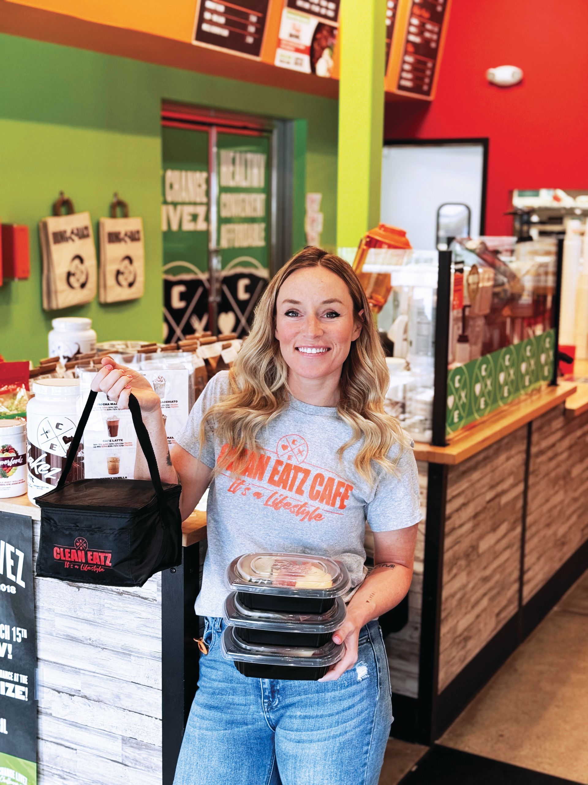 A woman in a gray t-shirt smiles while holding a black insulated bag and a stack of containers in a cafe.