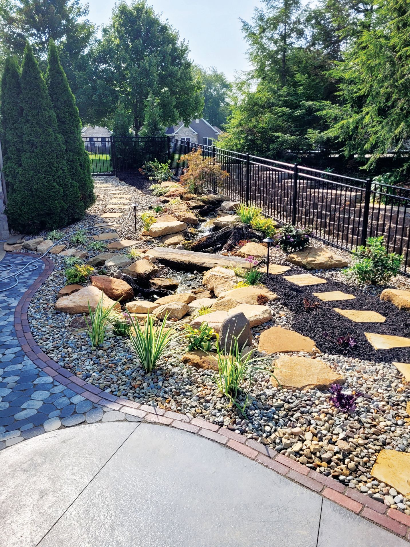 A backyard landscape featuring a stone-lined pond and stream, surrounded by decorative rocks, plants, and a paver patio.