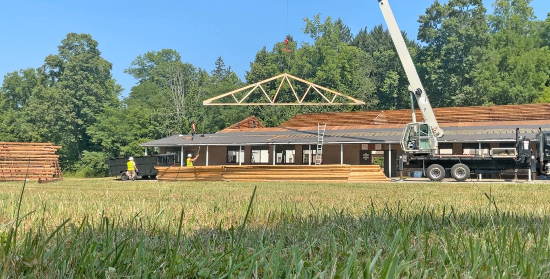 A construction crane lifts a large wooden roof truss above a low building in a grassy field on a sunny day.