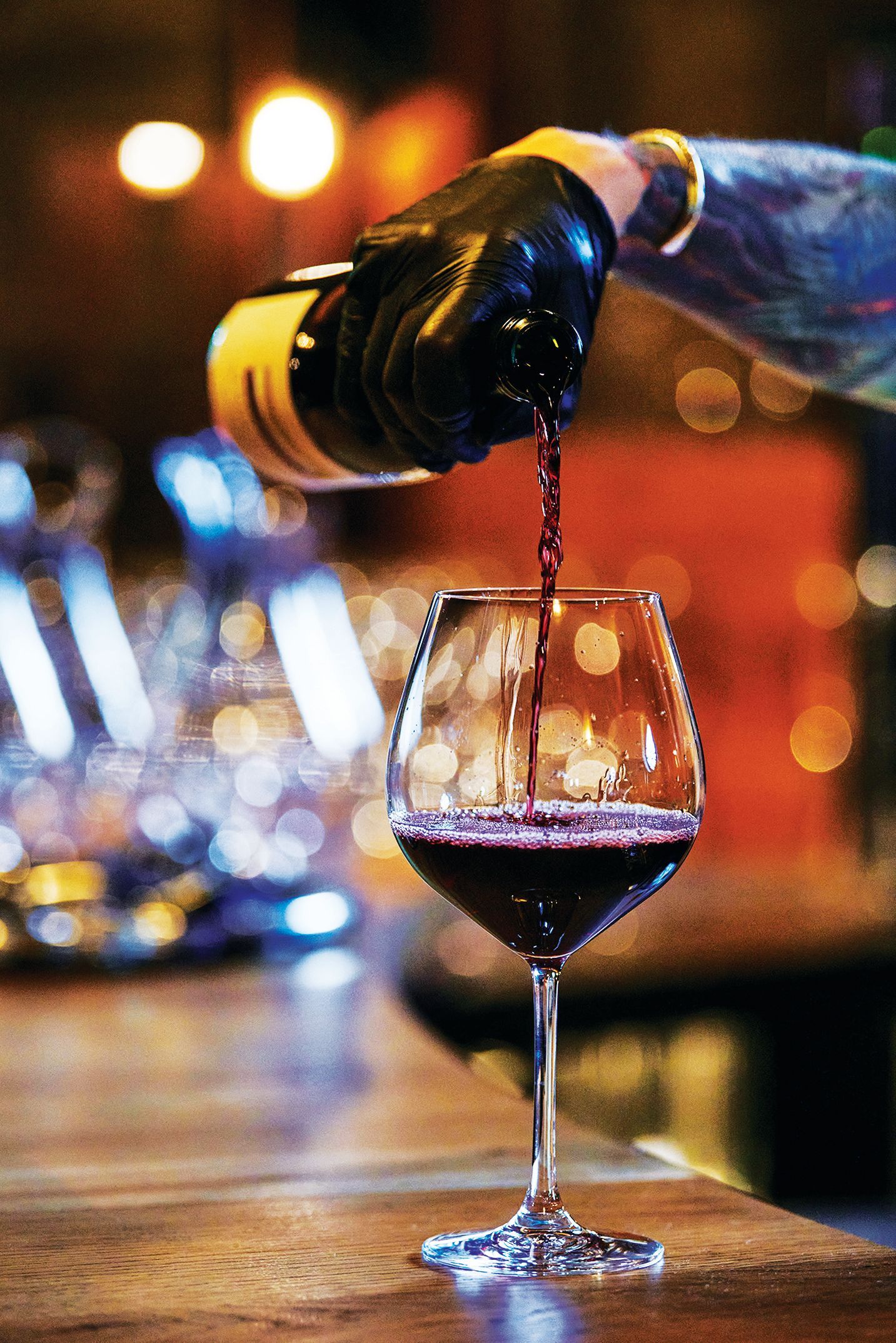 A hand wearing a black glove pours red wine from a bottle into a glass on a dimly lit bar counter.