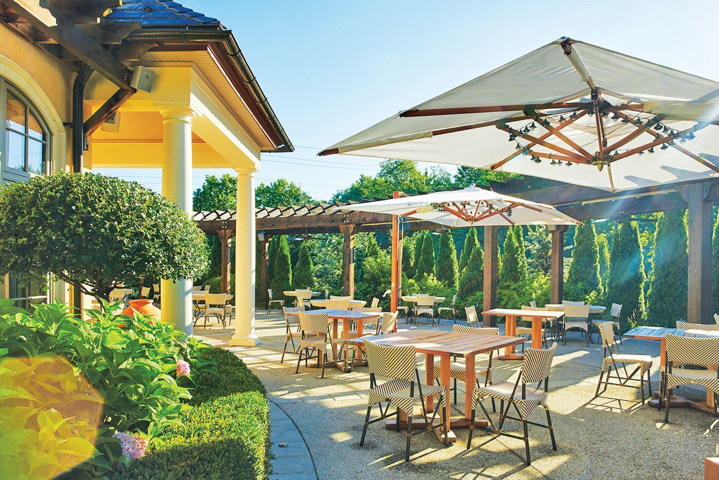 A sunlit outdoor restaurant patio with dining tables and umbrellas under a pergola beside a yellow building.
