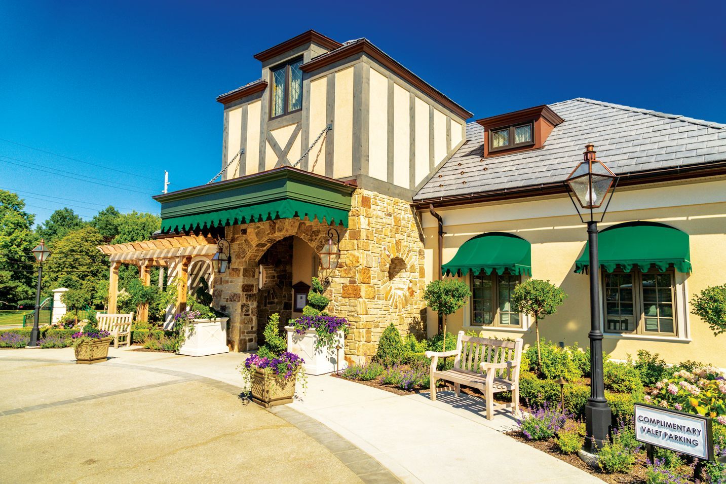 A charming, European-style building with stone accents, green awnings, a pergola, and a lamppost on a sunny day.