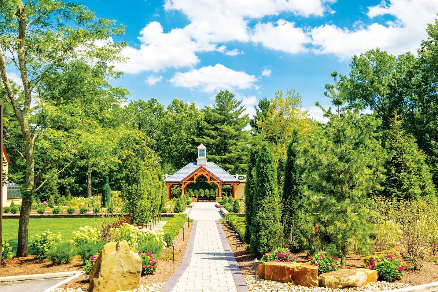 A paved stone path leads toward an open-air gazebo in a lush green garden under a blue sky with scattered clouds.