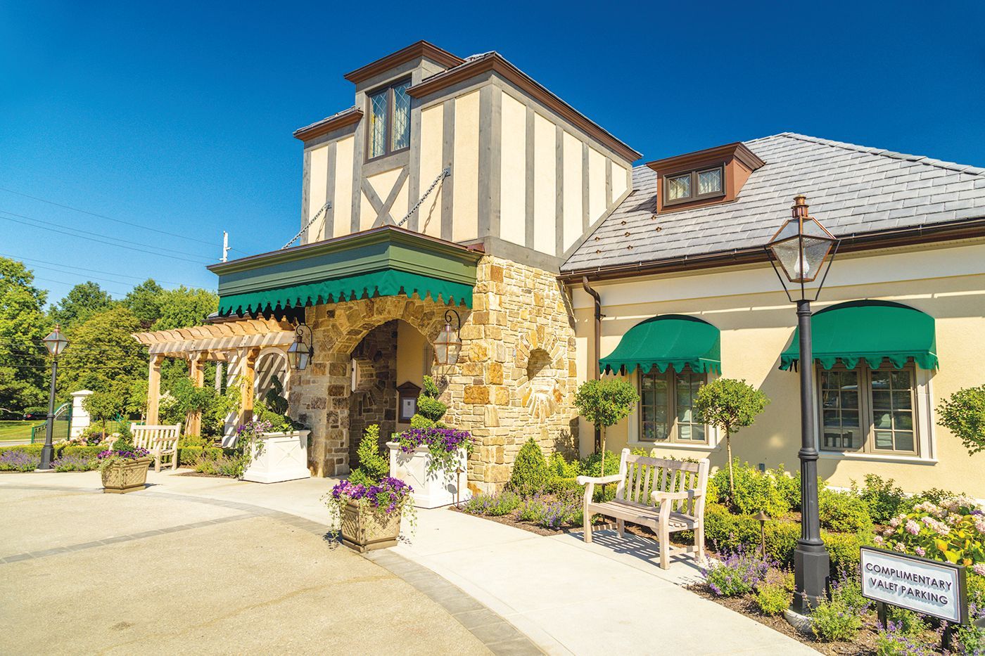 A charming stone building with a green awning, arched entrance, and a landscaped garden under a clear blue sky.