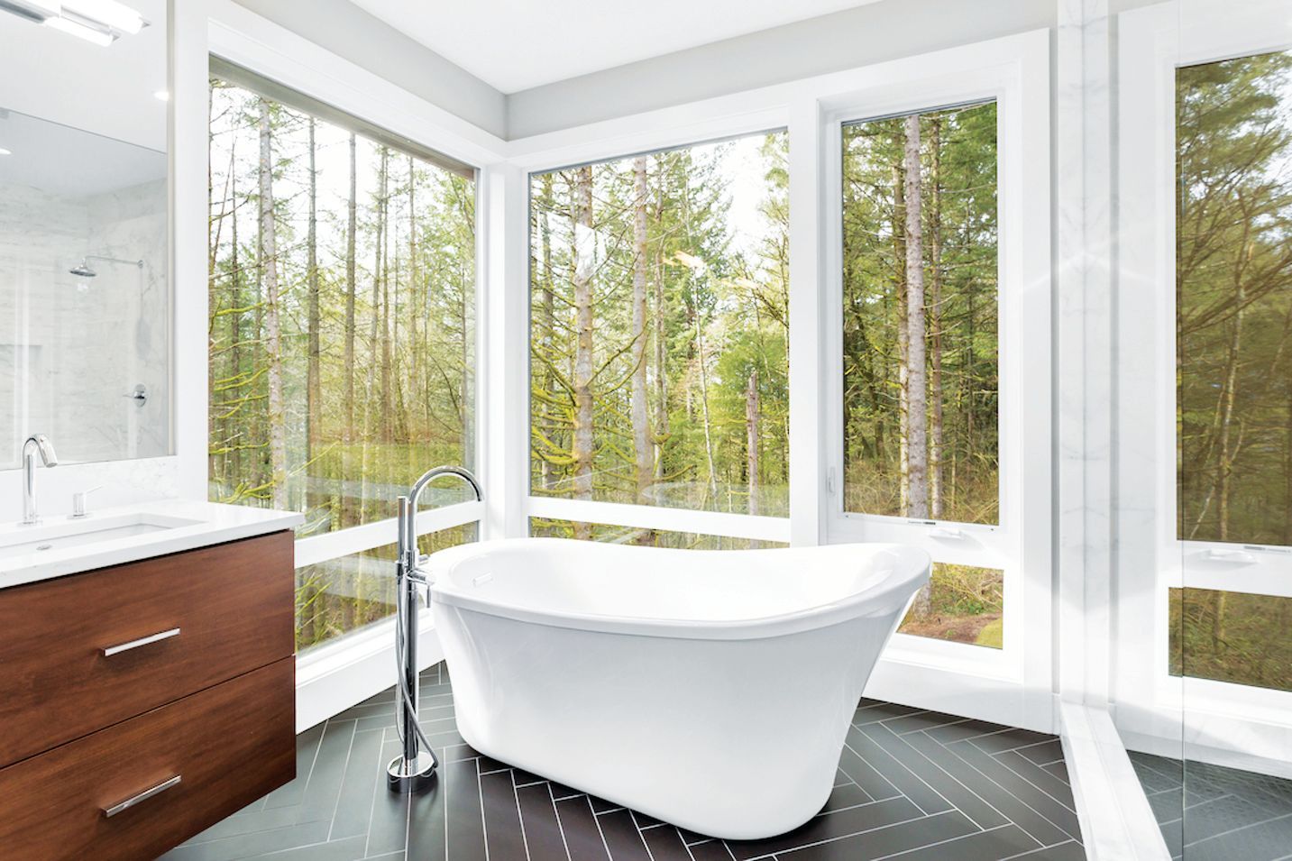 A modern bathroom with a white freestanding tub, a wooden vanity, and large windows overlooking a forest.
