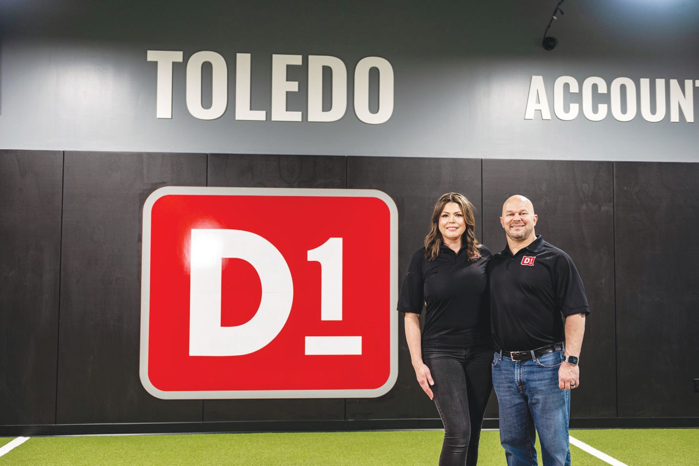 Two people stand side-by-side wearing black shirts in front of a red D1 logo on a wall labeled 