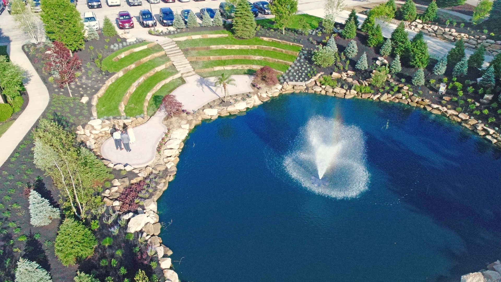An aerial view of a pond with a central fountain, adjacent to a terraced, grassy amphitheater and a parking lot.