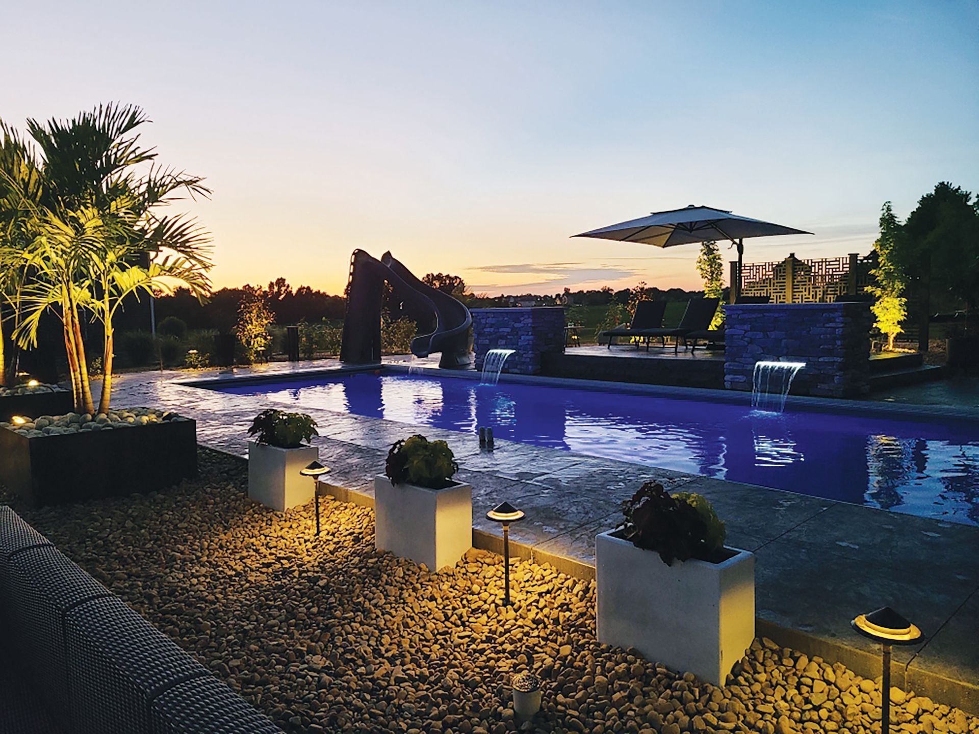 A backyard swimming pool at sunset, featuring a water slide, fountains, stone accents, planters, and glowing path lights.