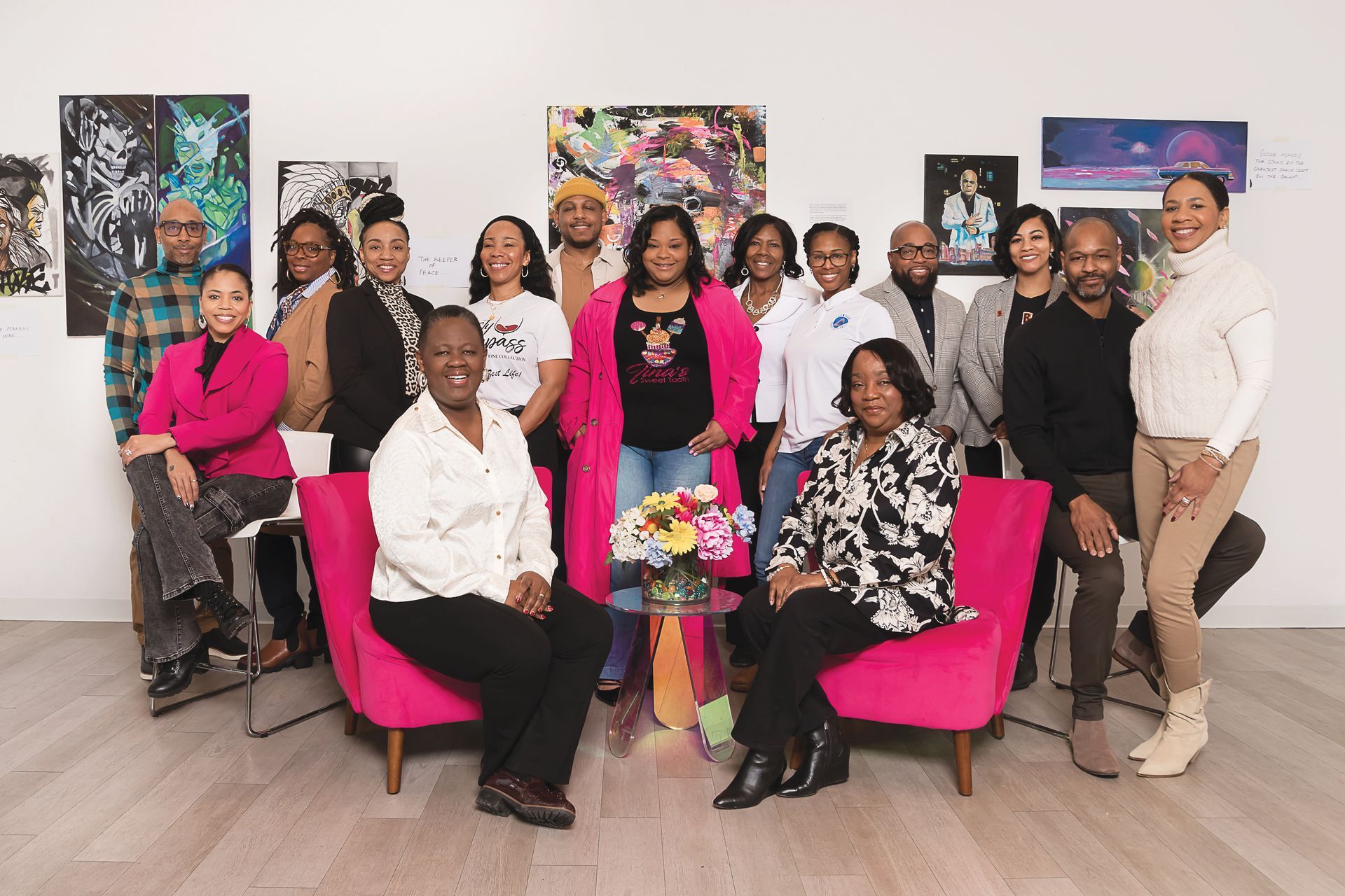 A group of people poses in an art gallery, with two people seated in bright pink chairs in front of art displays.
