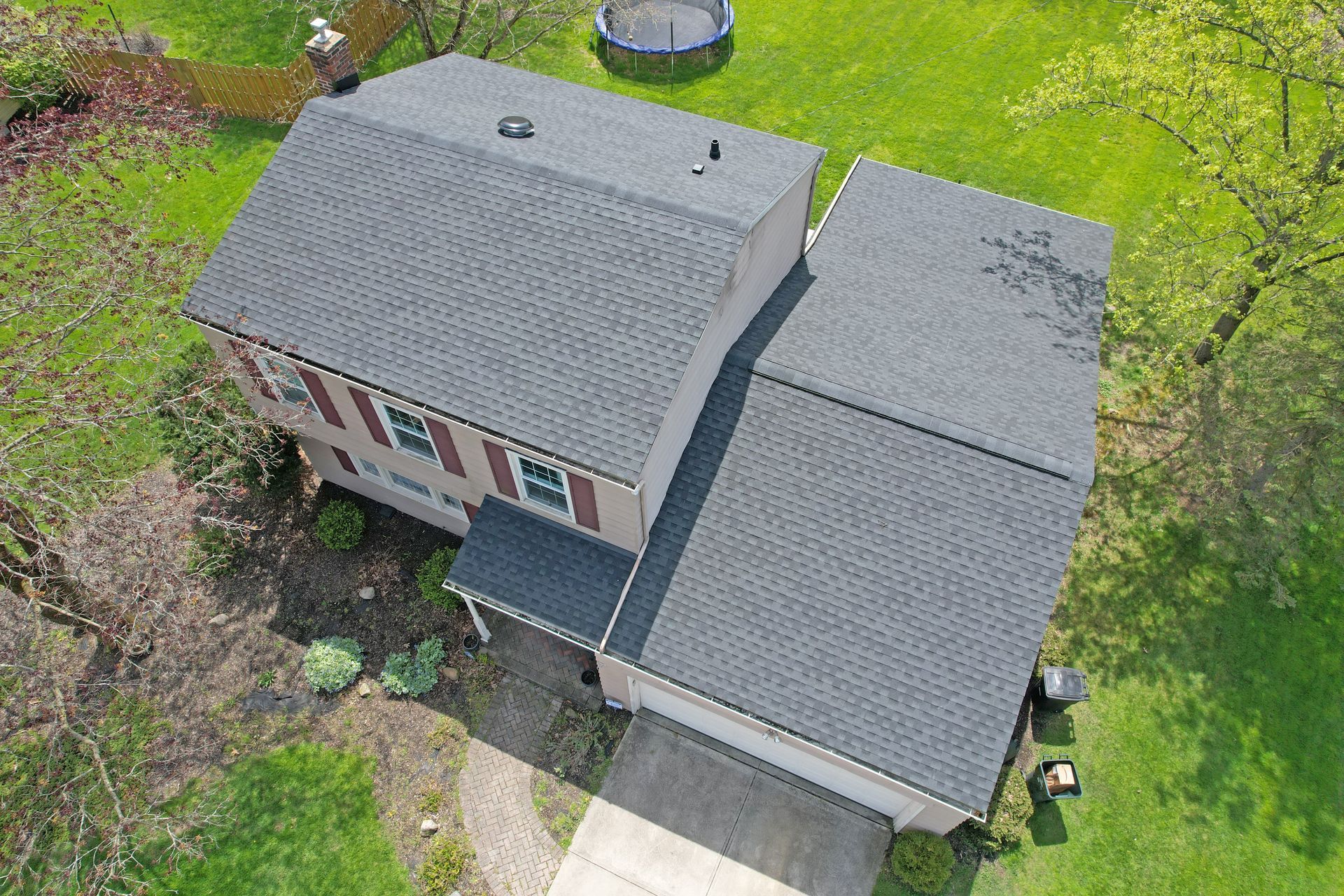An aerial view of a two-story house with a grey asphalt roof, surrounded by a green lawn and trees.