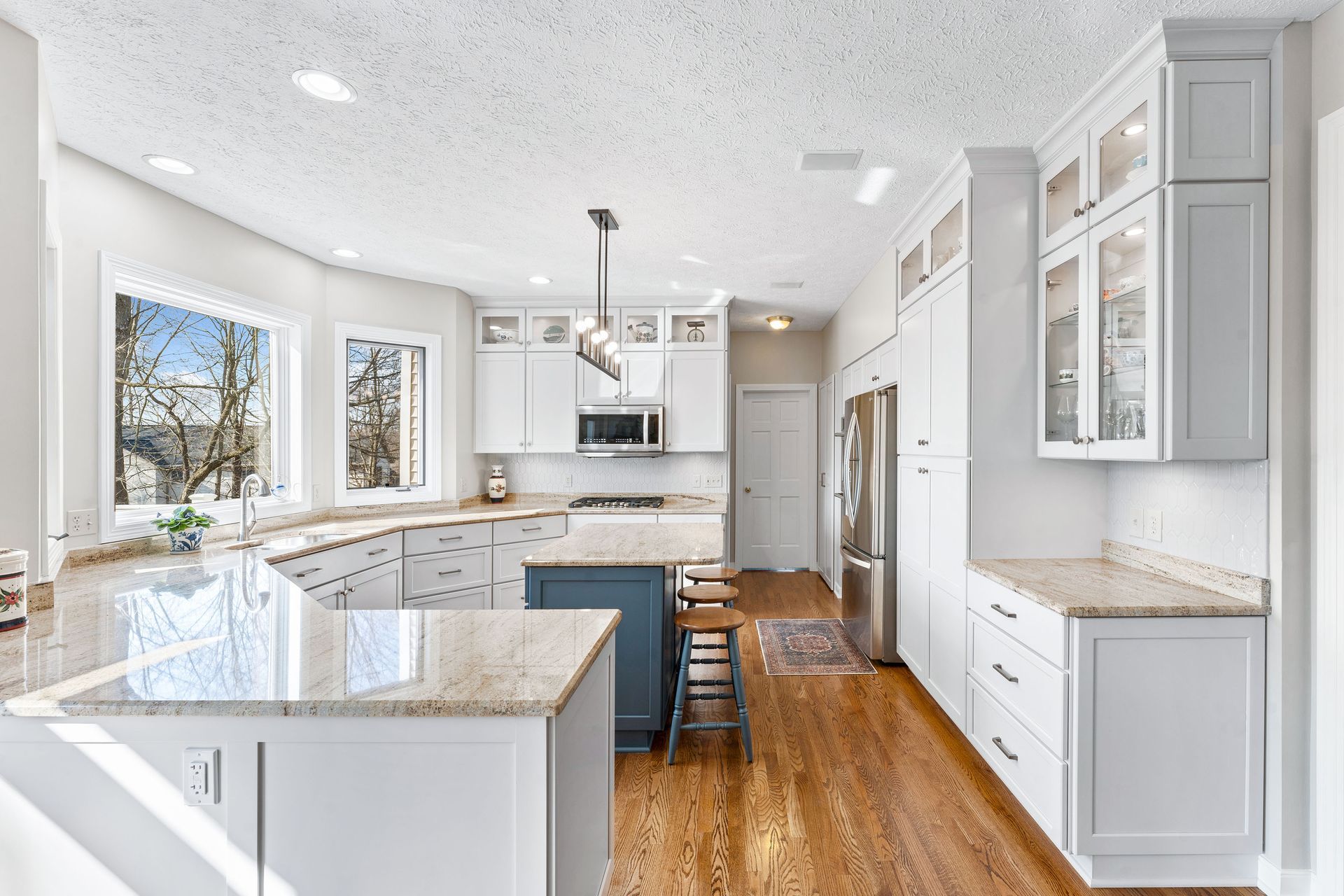 A modern white kitchen featuring an L-shaped granite counter, a blue island with wooden stools, and hardwood floors.