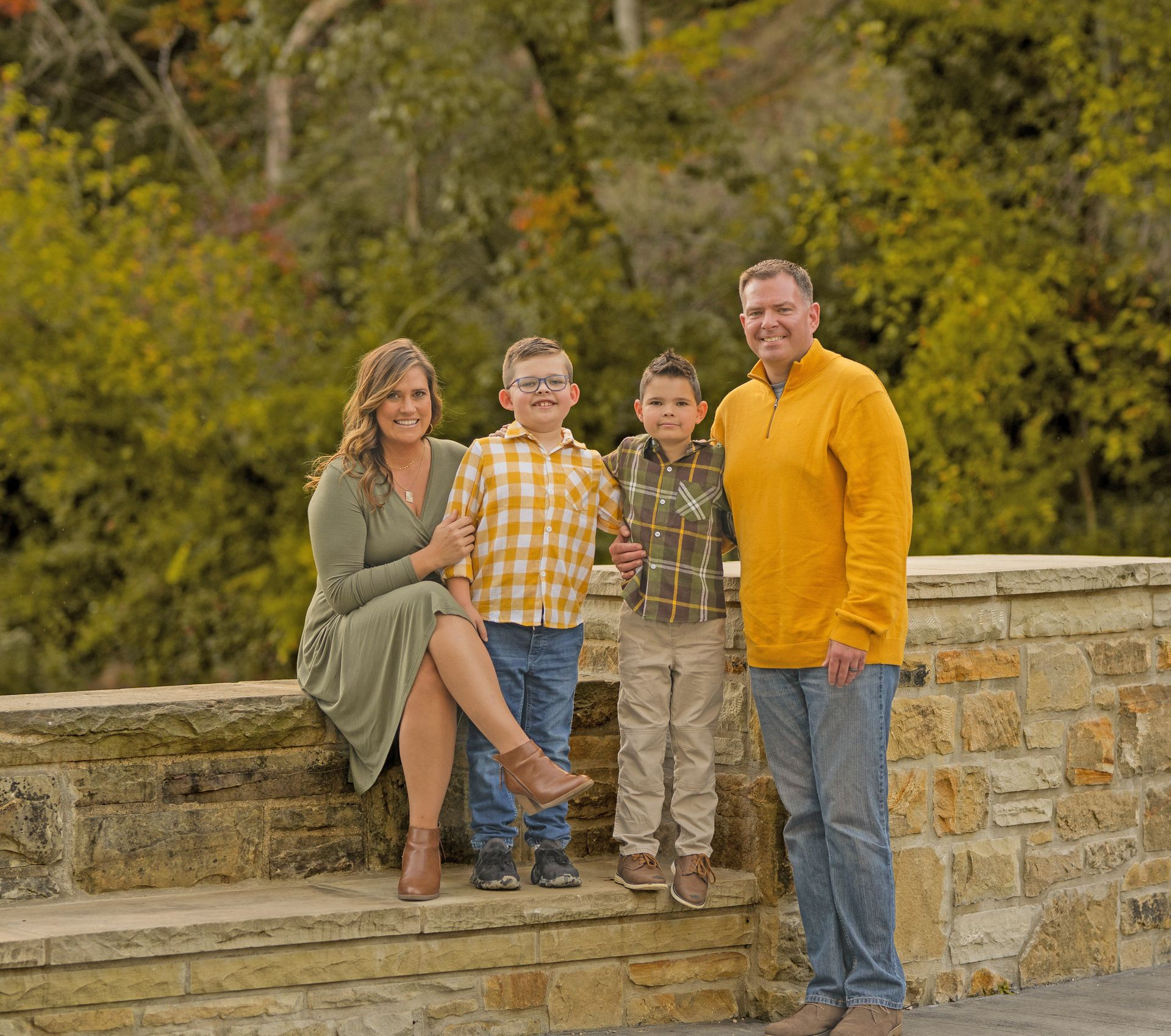 A family of four poses together outdoors on a stone wall in front of trees during autumn.