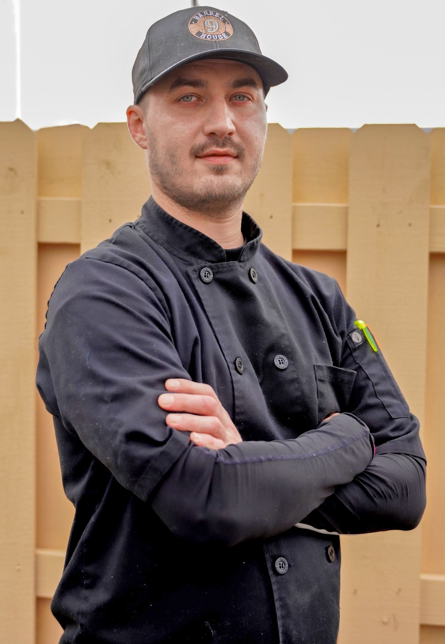 A person wearing a black chef’s uniform and baseball cap, standing with arms crossed in front of a tan wooden fence.