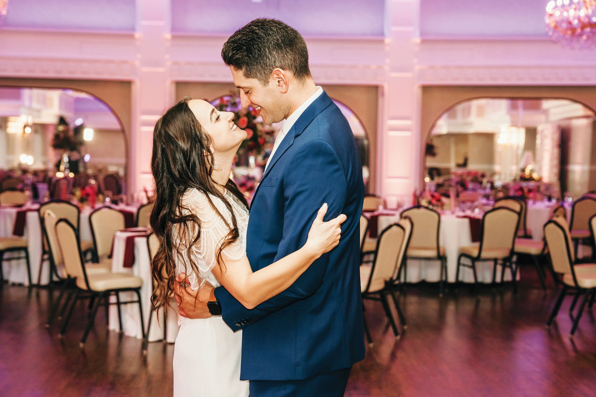 A couple embraces during a slow dance in a warmly lit, elegant ballroom featuring round tables and decorative chairs.