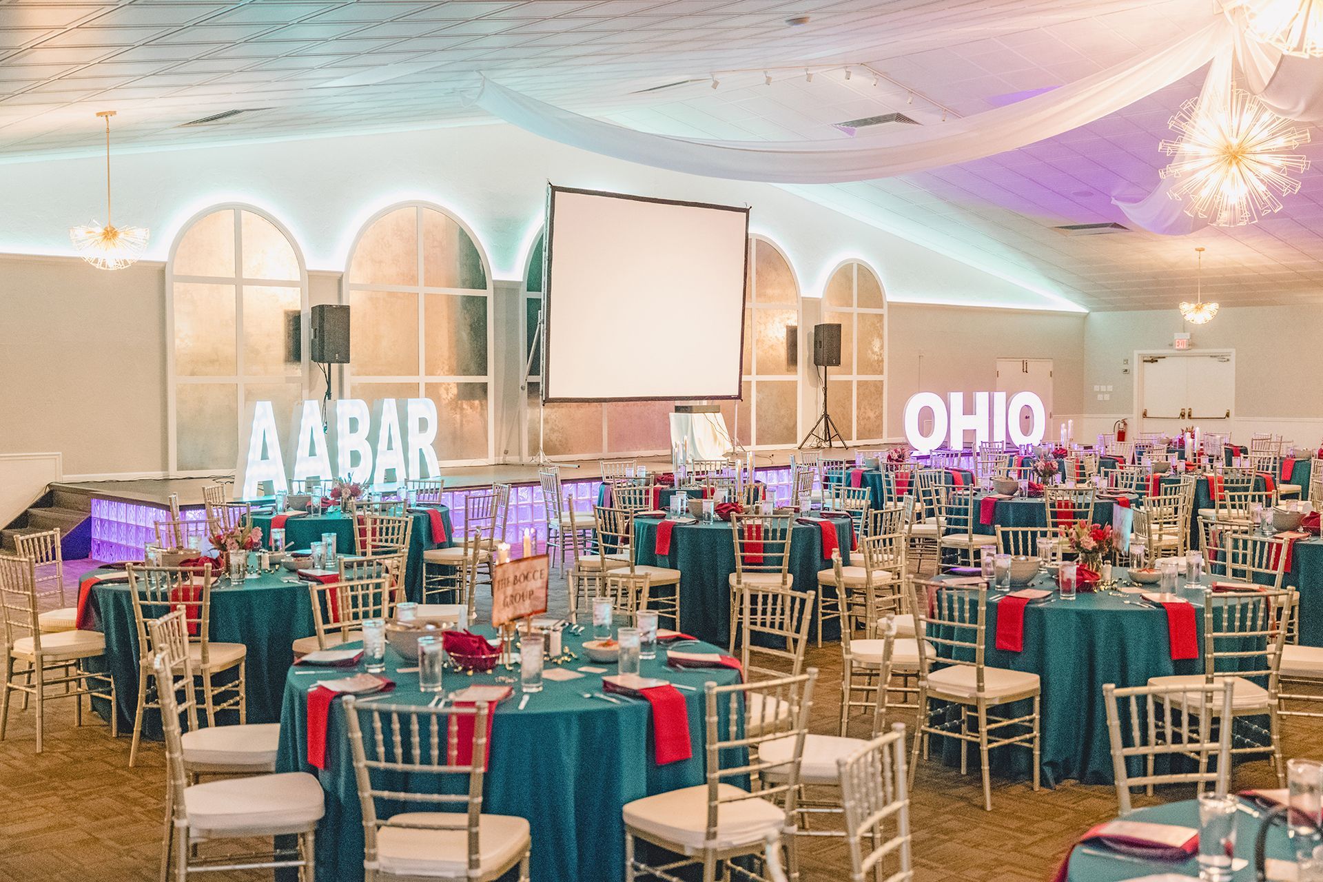 A brightly lit banquet hall set for a wedding with teal tablecloths, gold chairs, and glowing signs reading AABAR and OHIO.