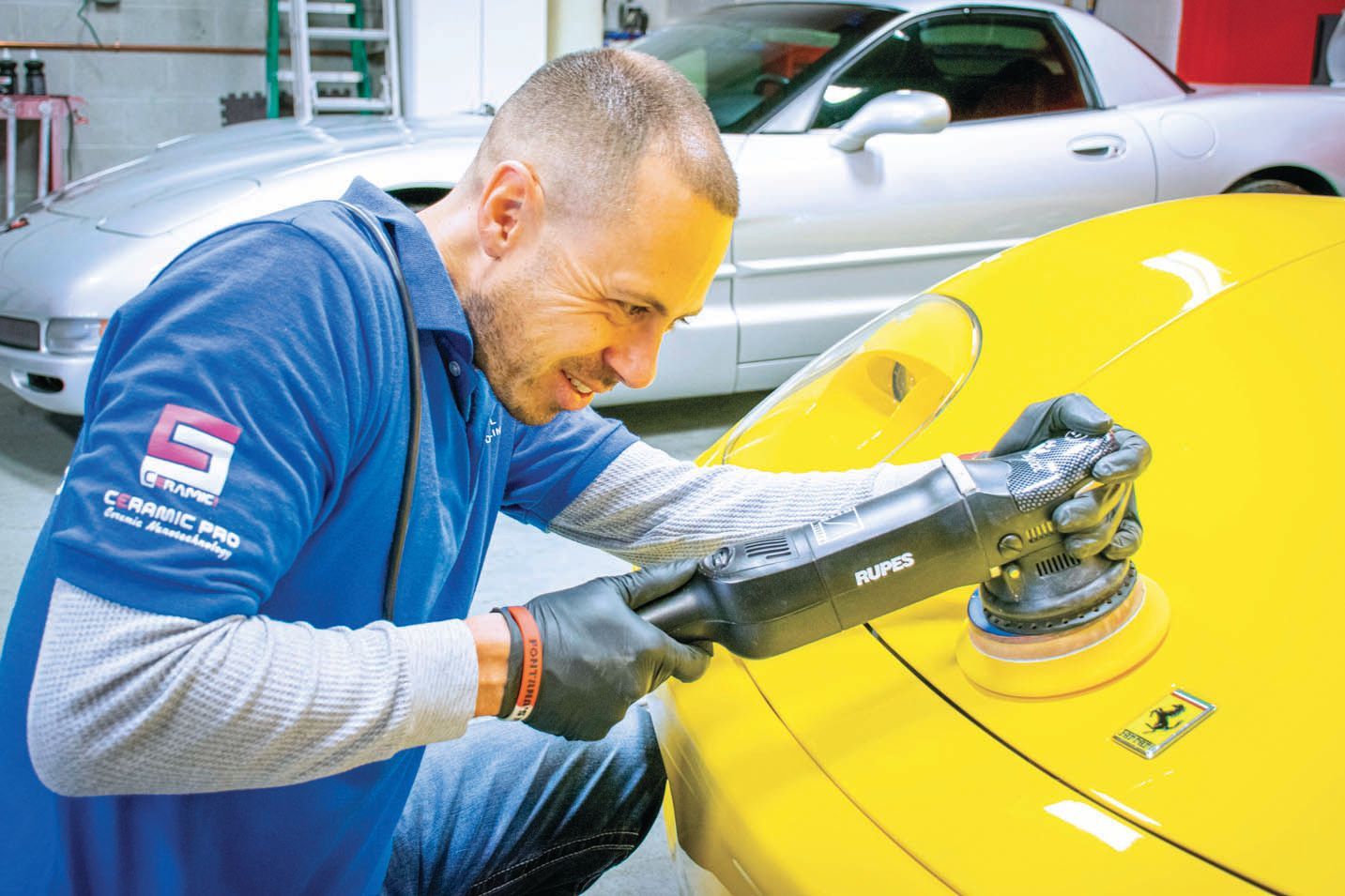 A person in a blue shirt uses a rotary polisher on the yellow hood of a Ferrari in a professional automotive garage.