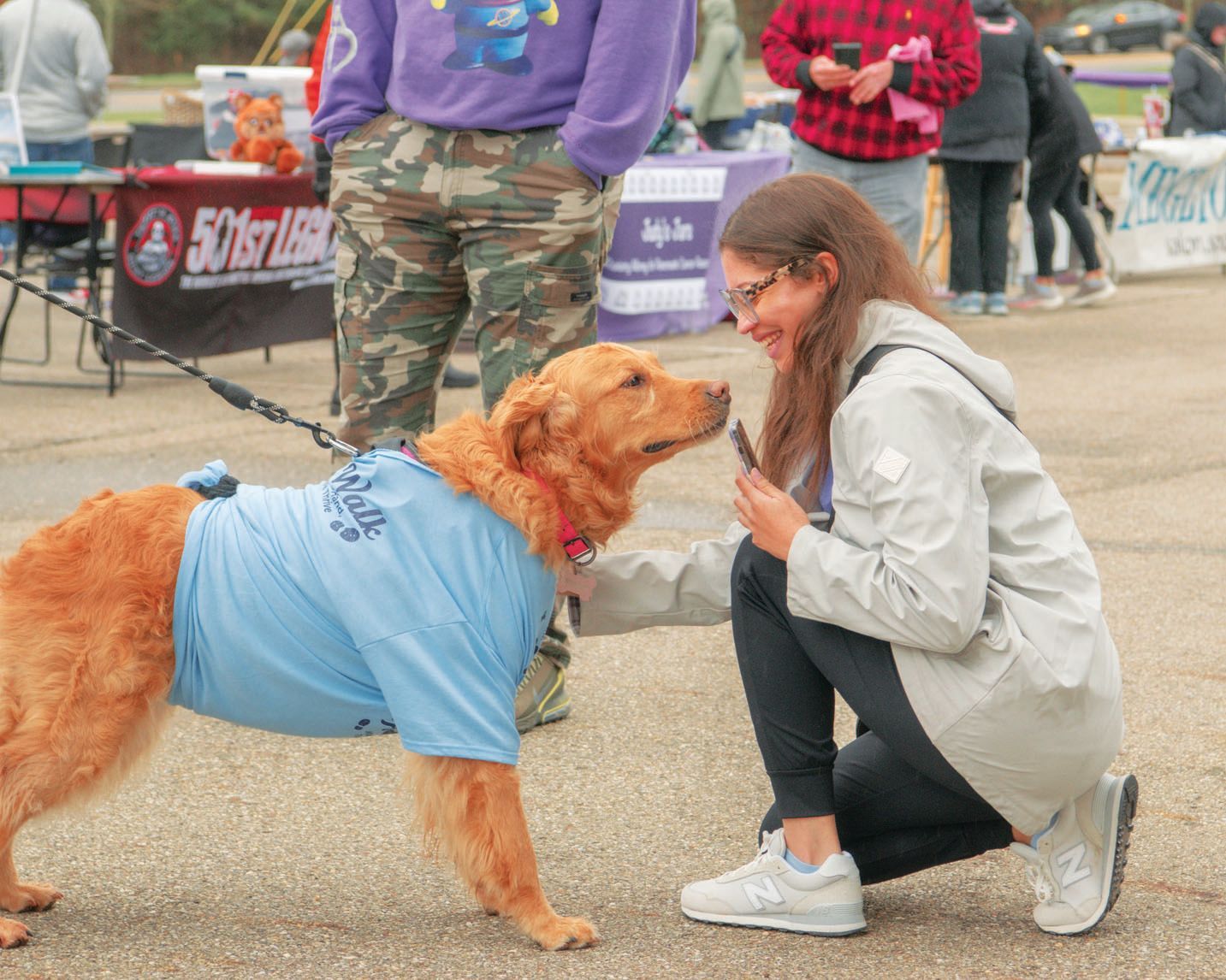 A person kneels to offer a treat to a golden retriever wearing a blue t-shirt outdoors at a community event.
