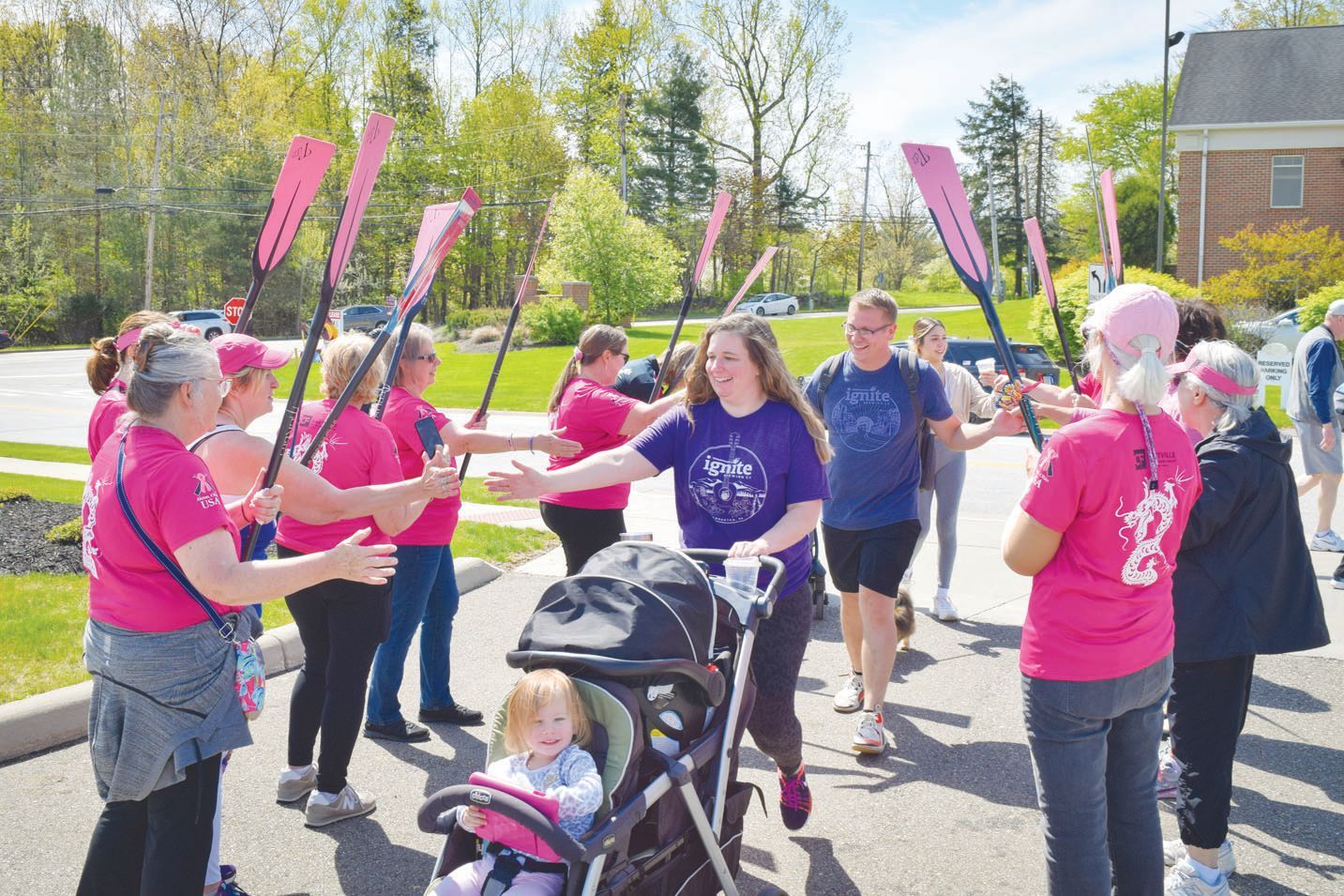 A group in matching pink shirts holds pink paddles, forming an honor guard for people walking past on a sunny day outdoors.