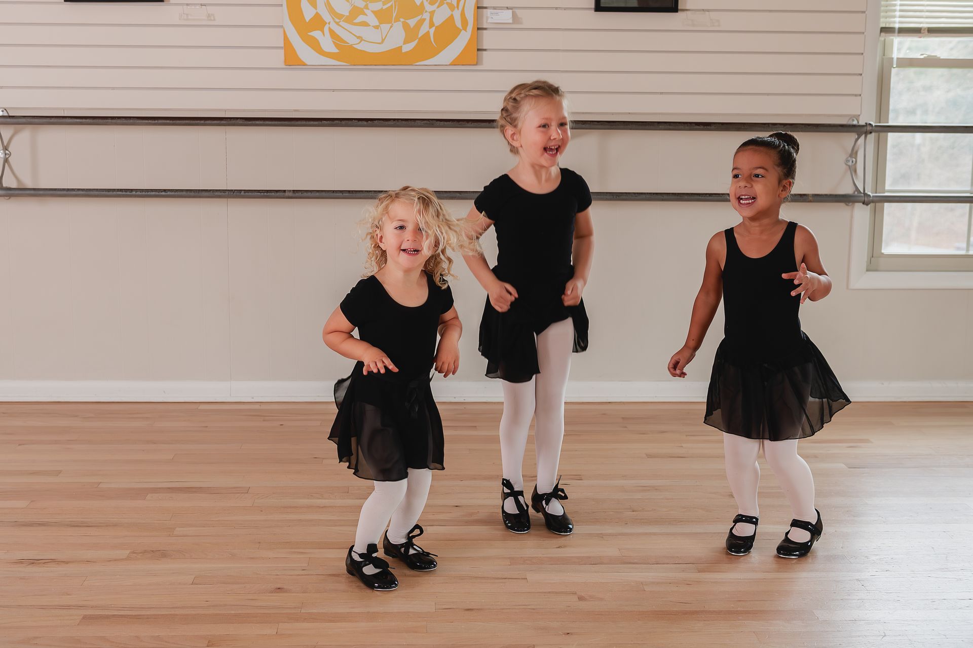 Three children in black leotards and dance skirts laughing while standing on a studio floor with ballet barres.