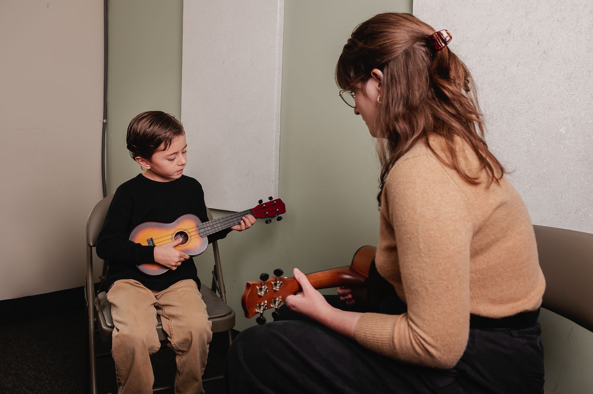 An instructor and a student sit in chairs, each holding a ukulele, during a music lesson in a room with neutral walls.