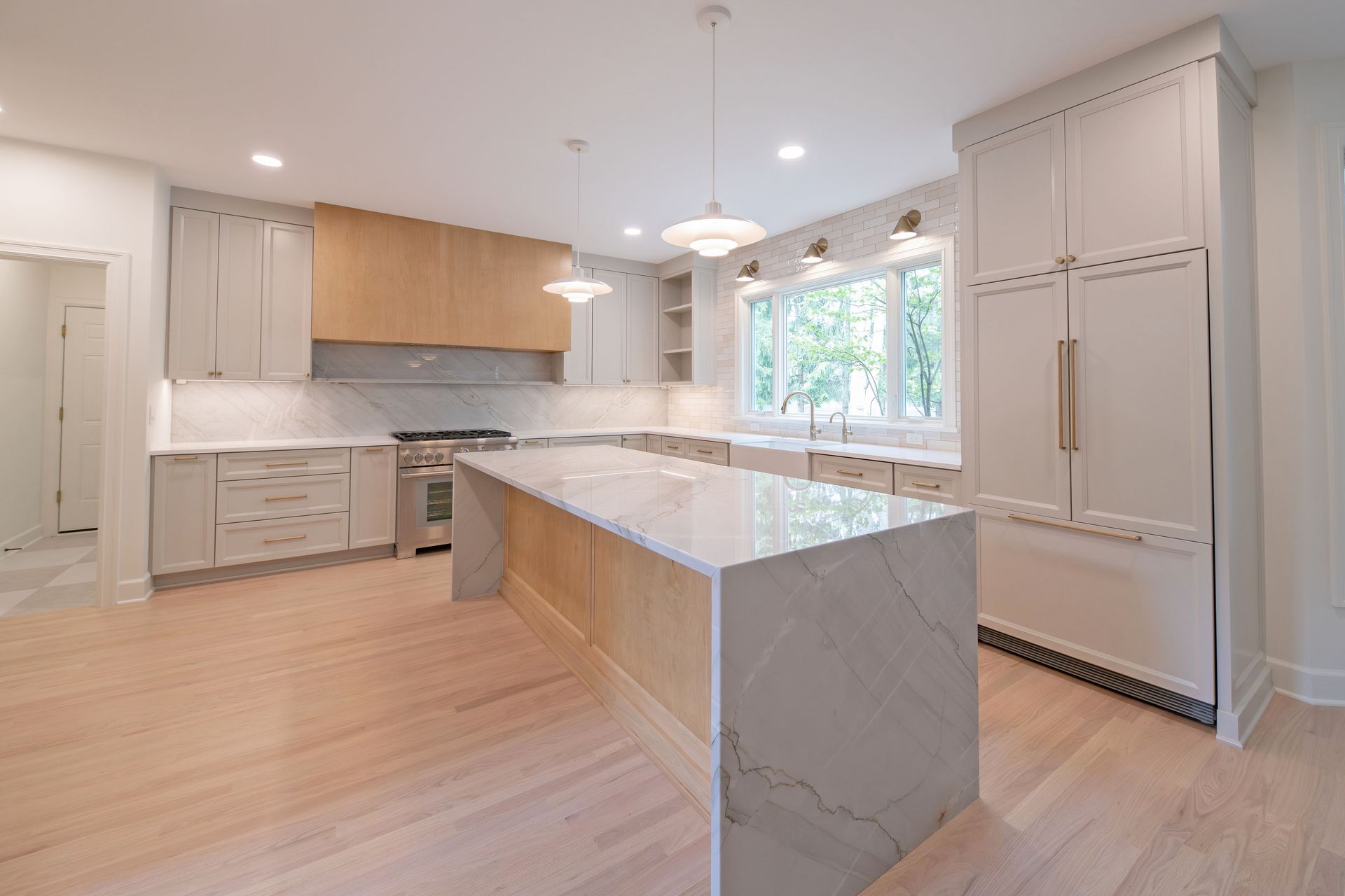 A modern kitchen with a large marble-topped island, light wood cabinets, stainless steel stove, and white floorboards.