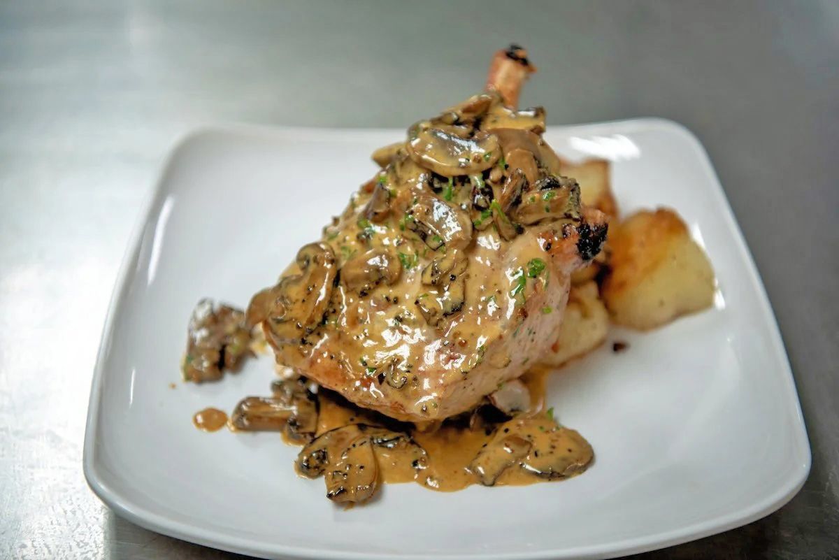 A seared pork chop topped with a creamy mushroom sauce, served with roasted potatoes on a square white plate.