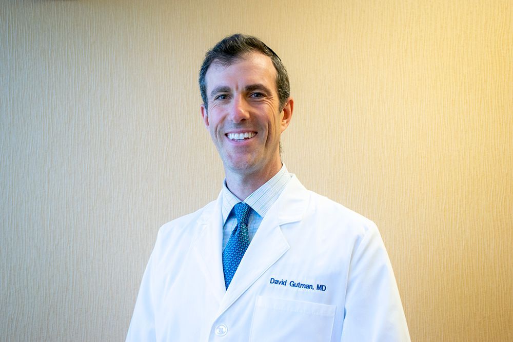 A smiling physician in a white lab coat and blue patterned tie, standing against a beige wall.
