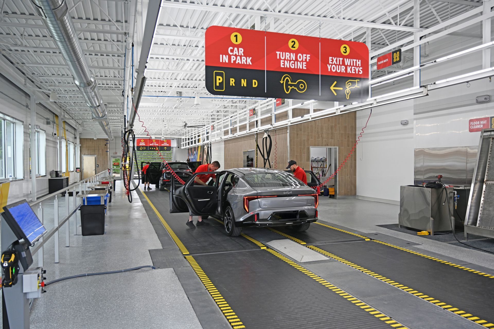 Employees guide a car into a bright, industrial car wash facility with instruction signage hanging overhead.