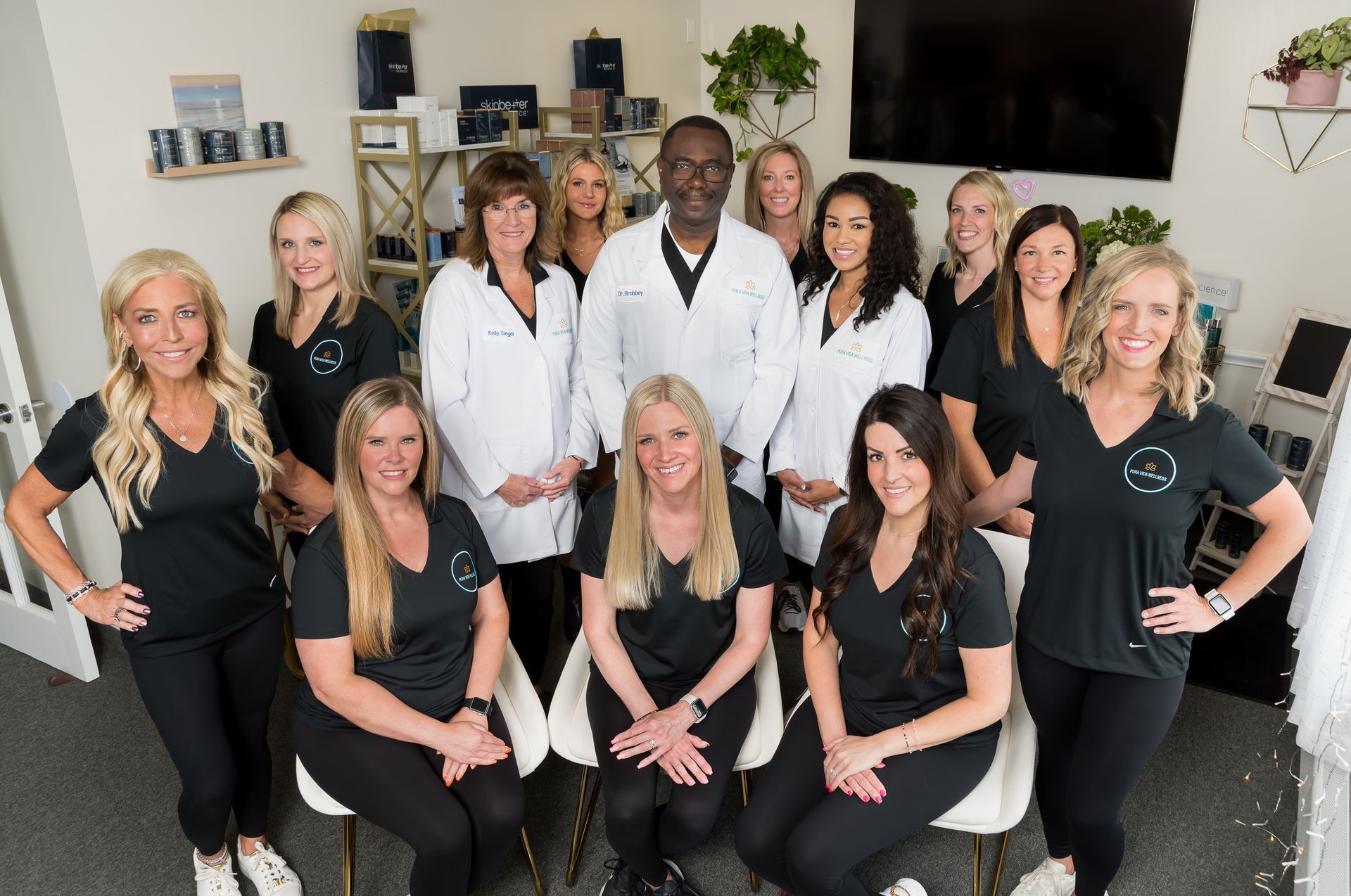 A group of thirteen people posed in a medical spa setting, wearing black tops and white coats, smiling for a group portrait.