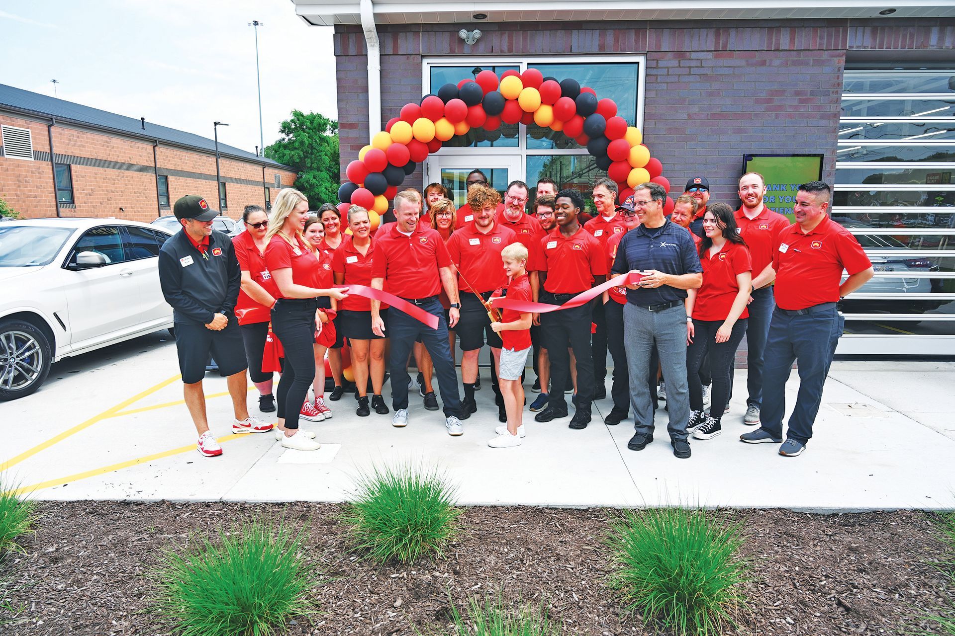 A group of people wearing red and black uniforms perform a ribbon-cutting ceremony in front of a building with balloons.