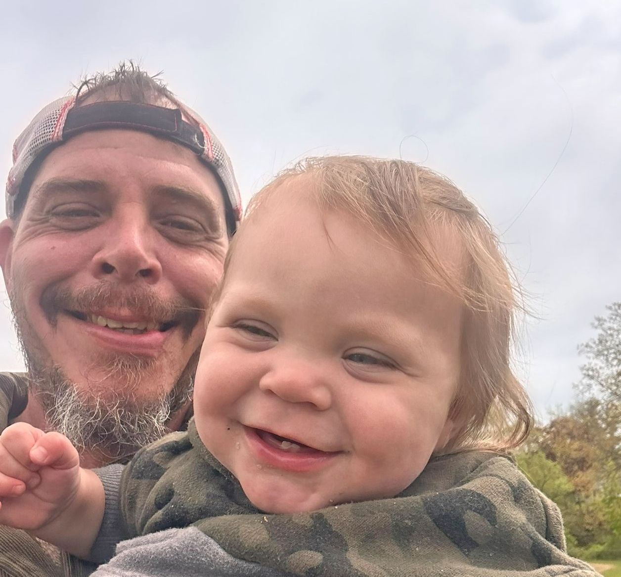 A smiling person wearing a baseball cap holds a smiling child in a camo-print shirt outdoors against a cloudy sky.