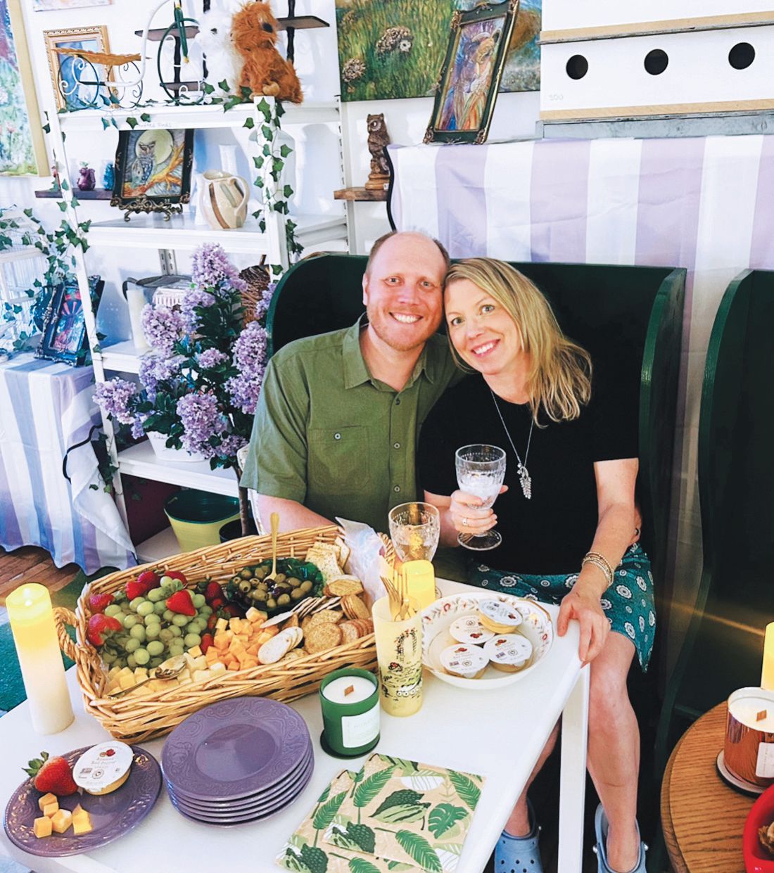 A couple smiles while sitting at a table with a basket of fruit, crackers, and snacks, holding glasses of drinks.