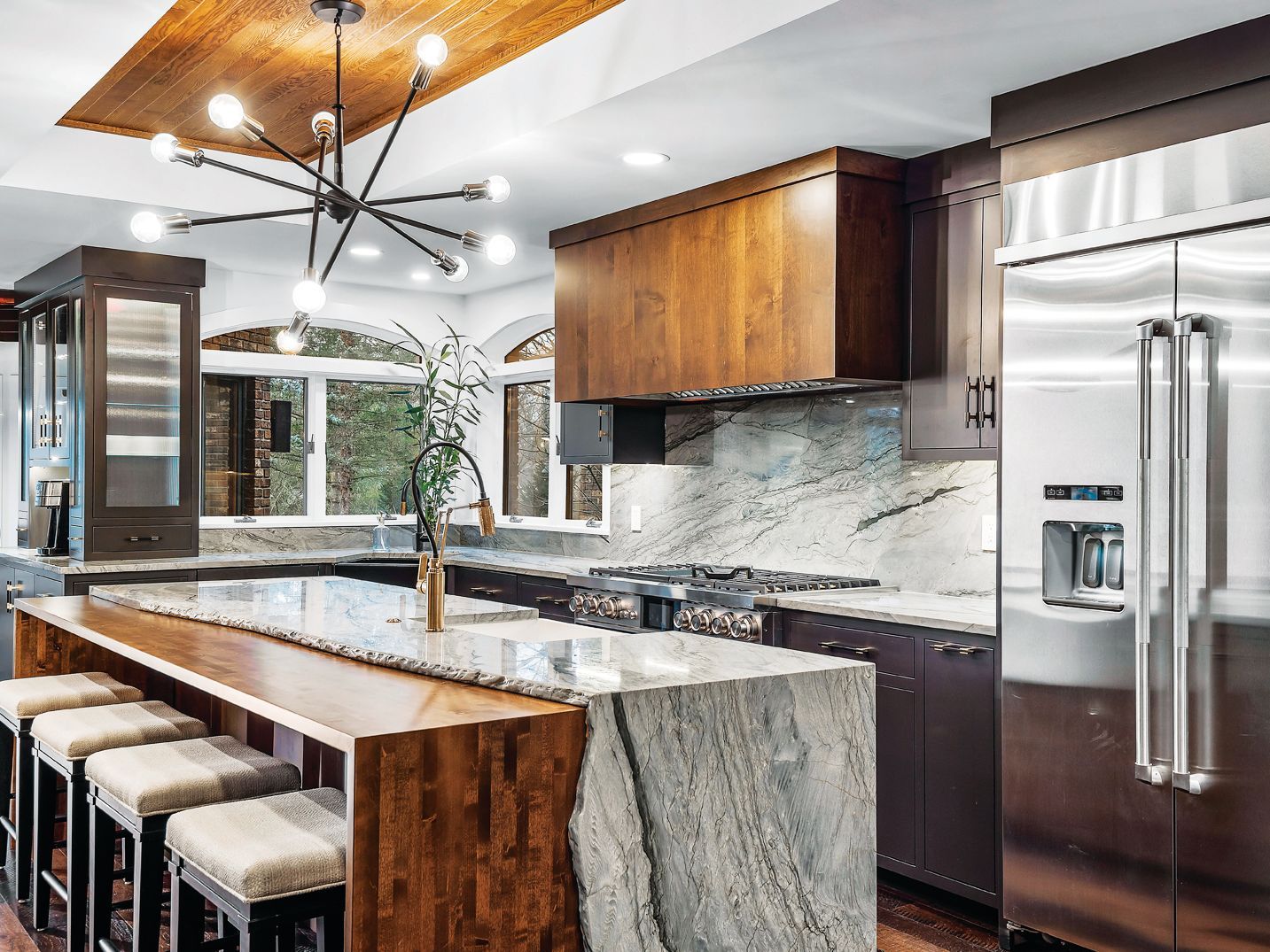 Modern kitchen with a granite island, breakfast bar stools, wooden cabinetry, and a stainless steel refrigerator.