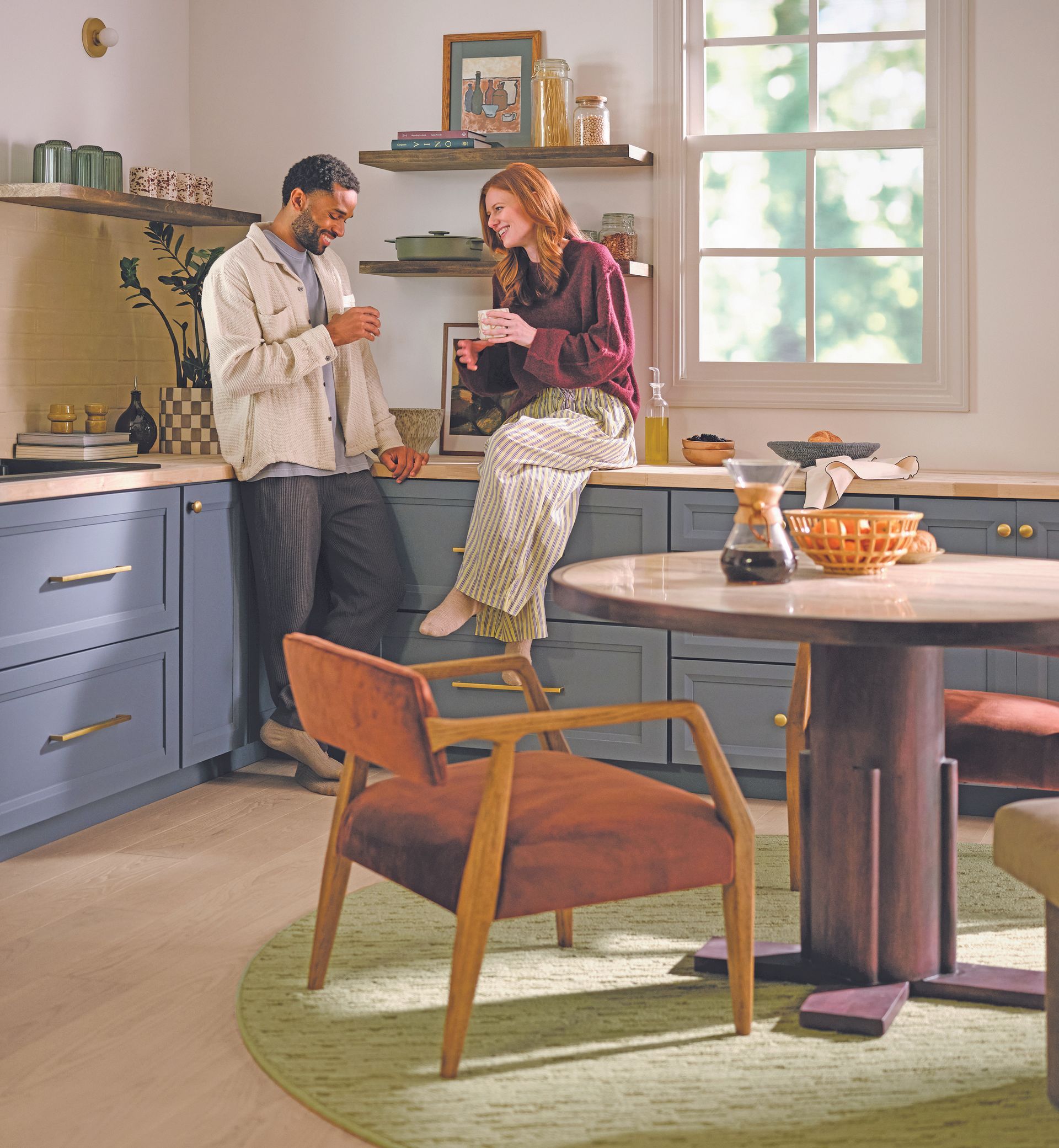A person standing and another sitting on a blue kitchen counter, laughing together in a sunlit, modern home interior.